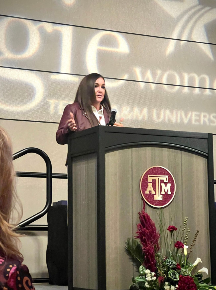 A woman speaking at a podium with the Texas A&M logo, with a large screen behind her displaying text related to Texas A&M University.