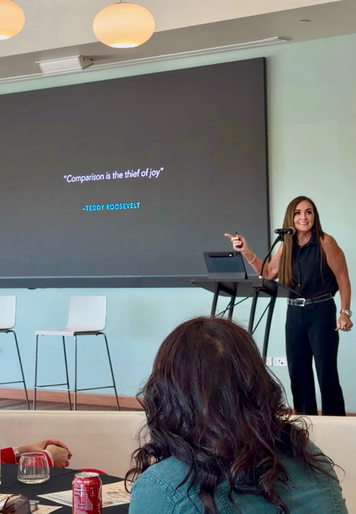 A woman giving a presentation in a conference room, standing at a podium with a microphone, with a large screen behind her displaying a quote by Teddy Roosevelt that says, 'Comparison is the thief of joy.'