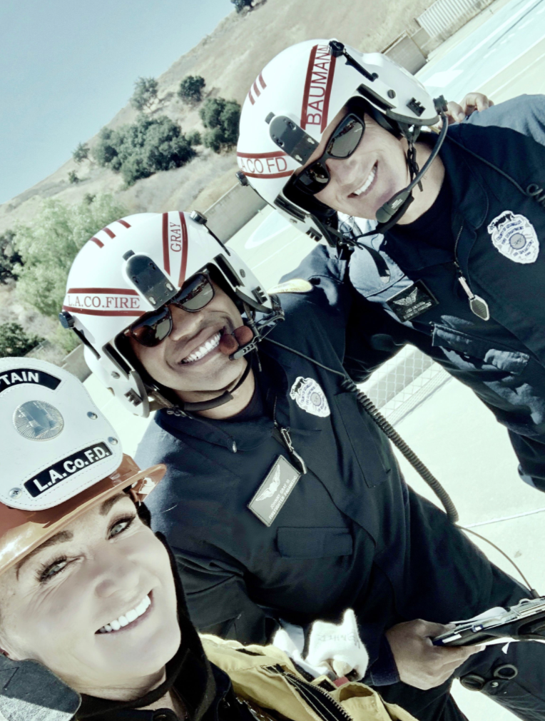 Three firefighters in uniform and helmets, smiling and taking a selfie outdoors on a sunny day.