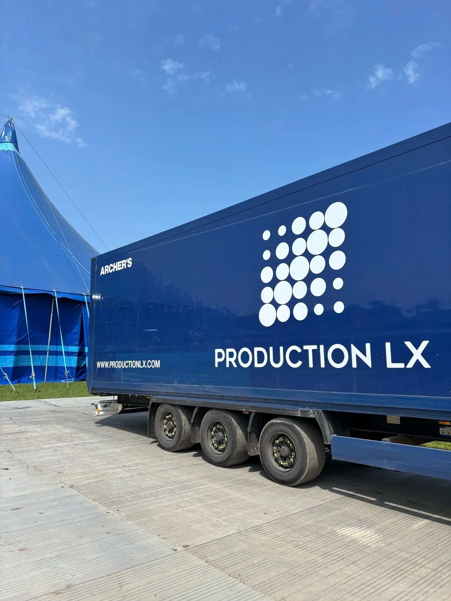 A blue truck with white text and logo, parked on a concrete surface, under a blue sky with some clouds.
