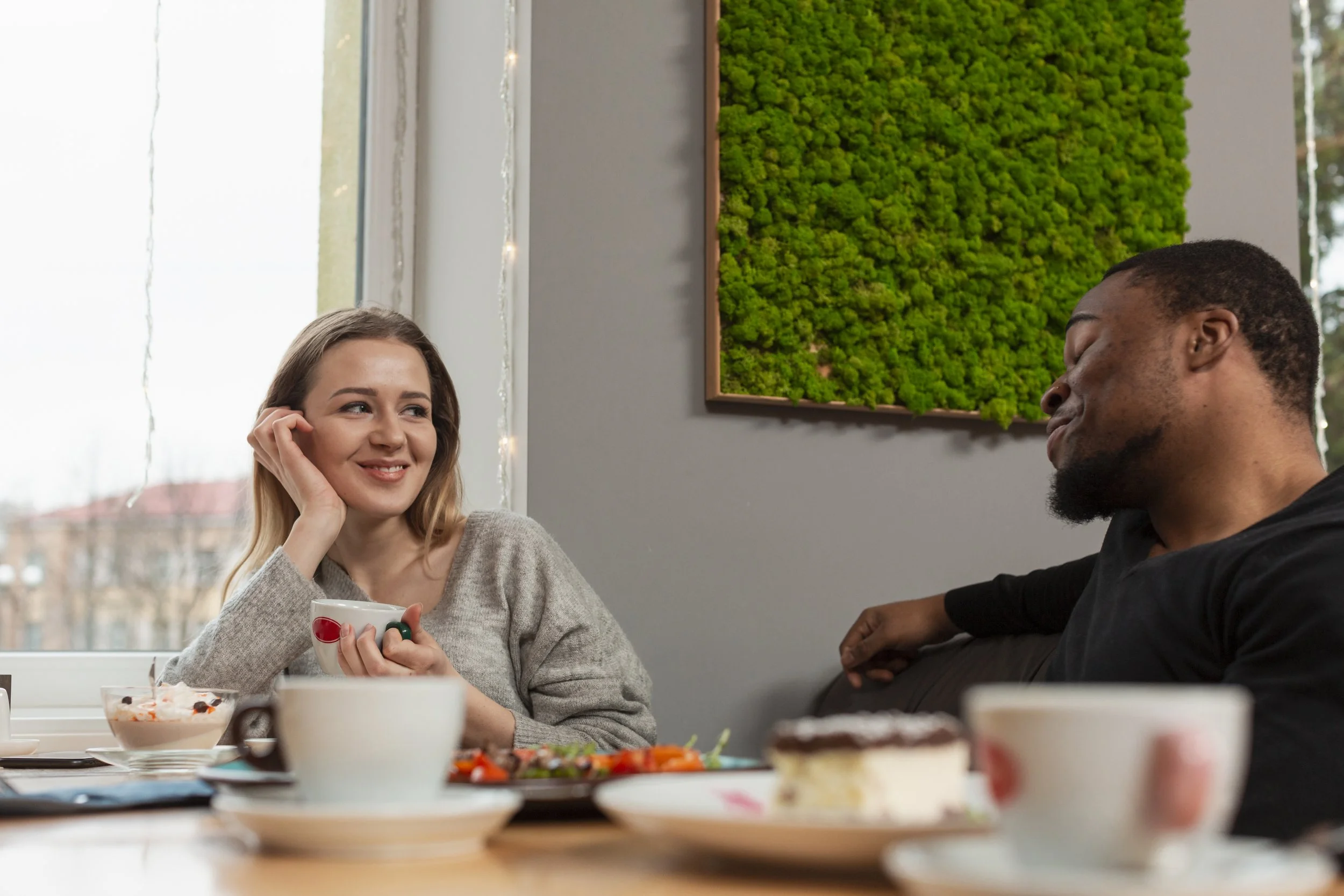 A woman and a man sitting at a table with desserts, coffee, and plates, enjoying a conversation in a cozy room with a green moss wall art and string lights in the background.