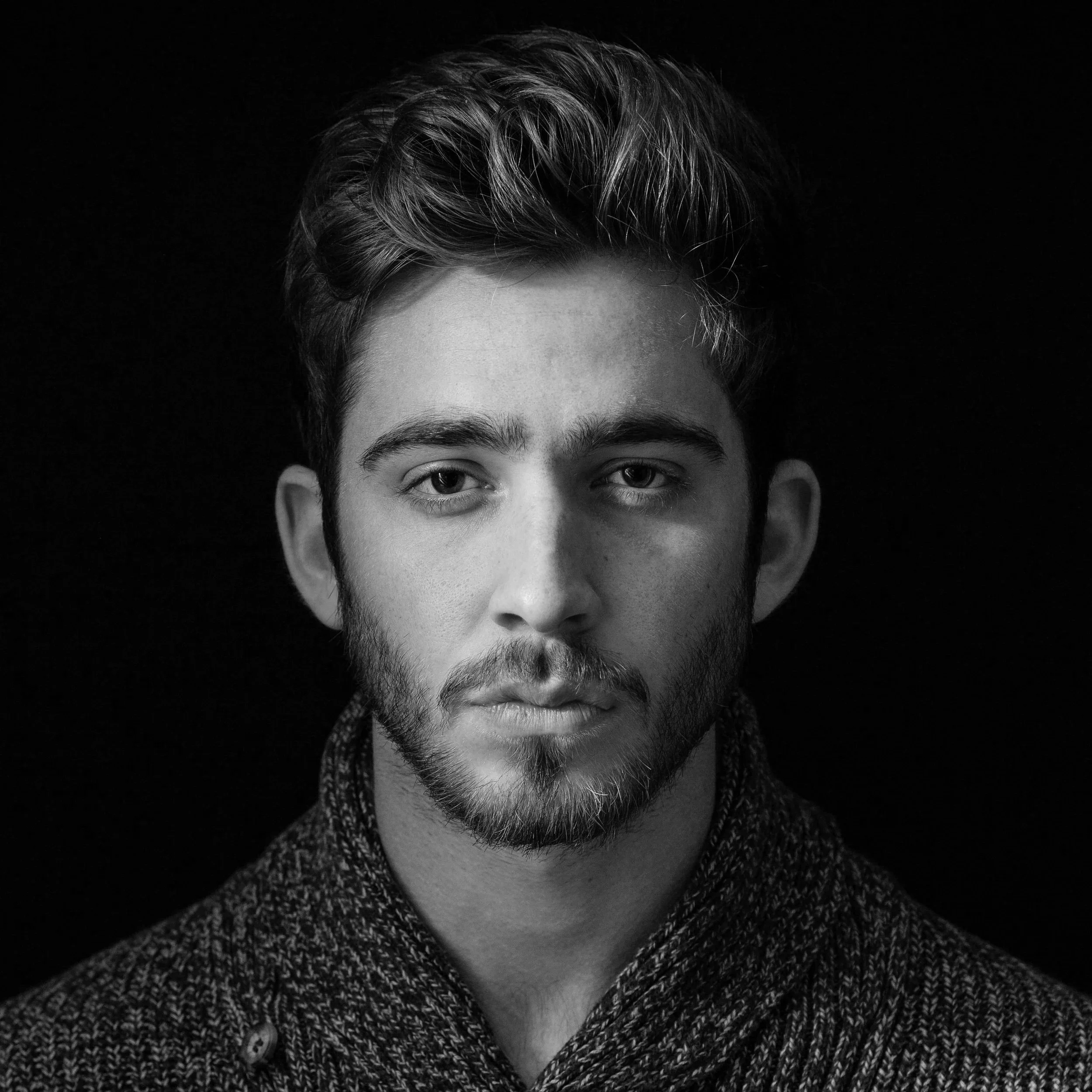 Close-up black-and-white portrait of a person with short, styled hair, mustache, and beard, wearing a textured coat, against a dark background.