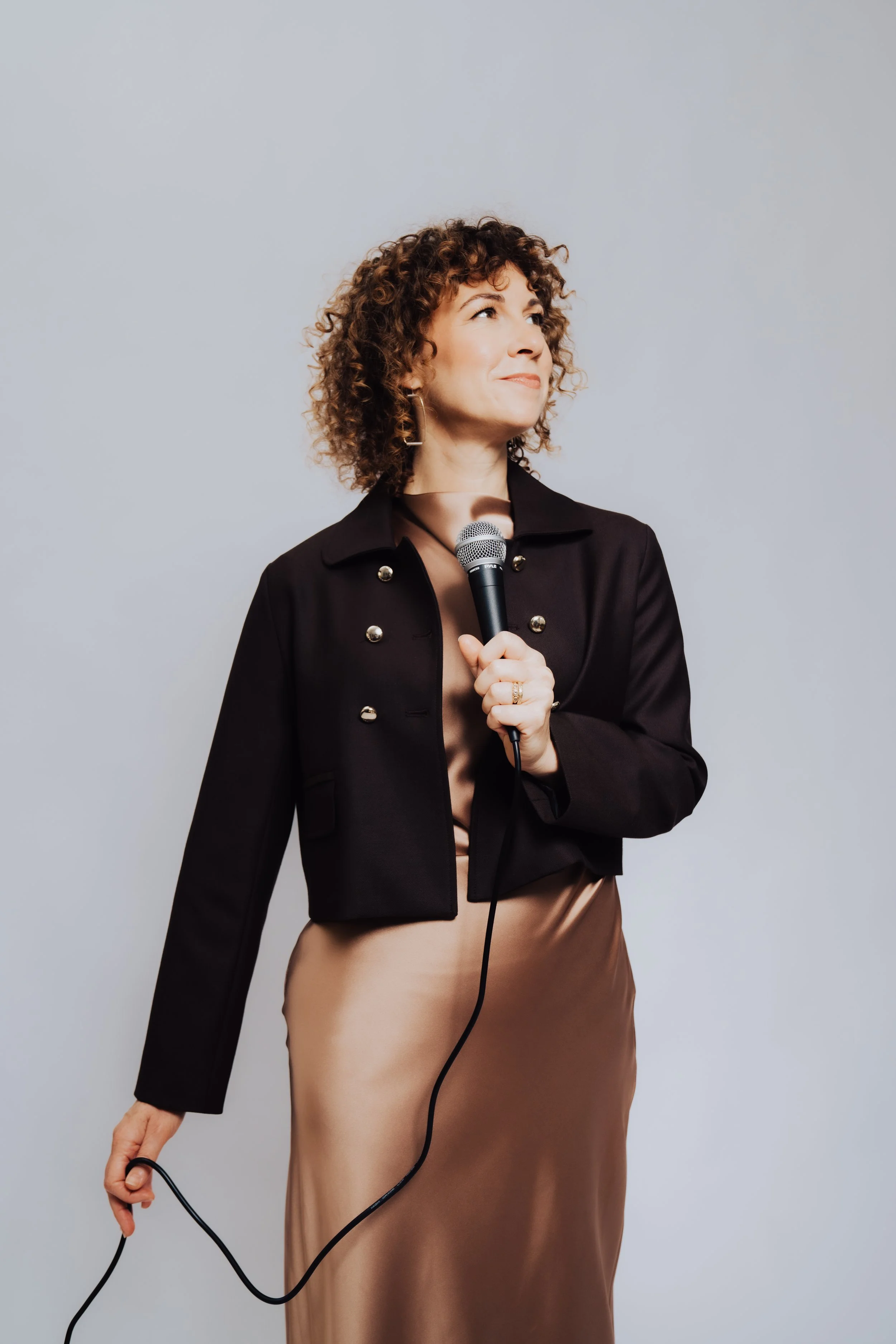 Woman with curly hair holding a microphone and a cord, dressed in a black blazer and brown satin skirt, standing against a plain gray background.