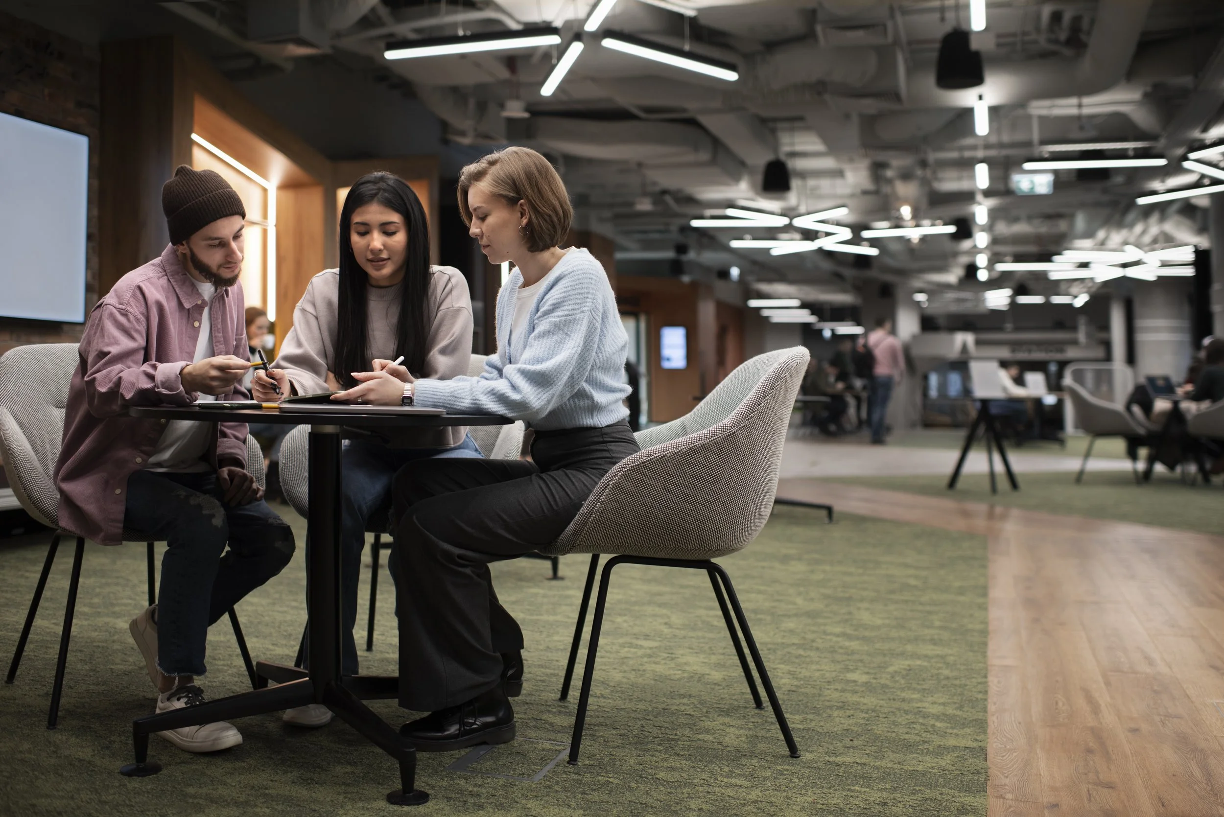 Three young women sitting at a round table in a modern office lounge, engaged in a discussion while looking at a tablet or phone. The background features other people working or walking in the open-plan office space with contemporary lighting and decor.