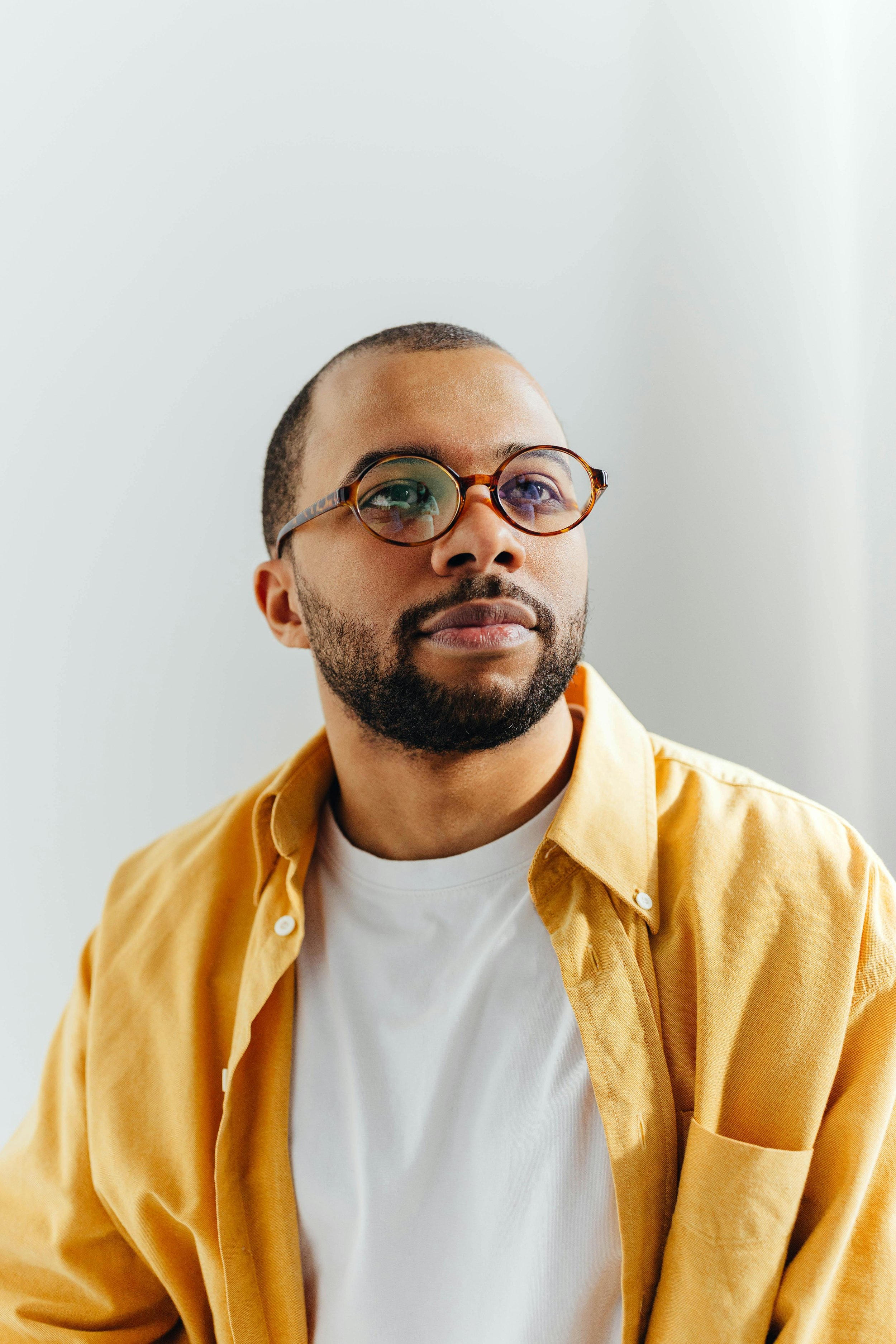 Close-up of a man wearing round glasses, a white T-shirt, and a yellow button-up shirt, against a plain white background.