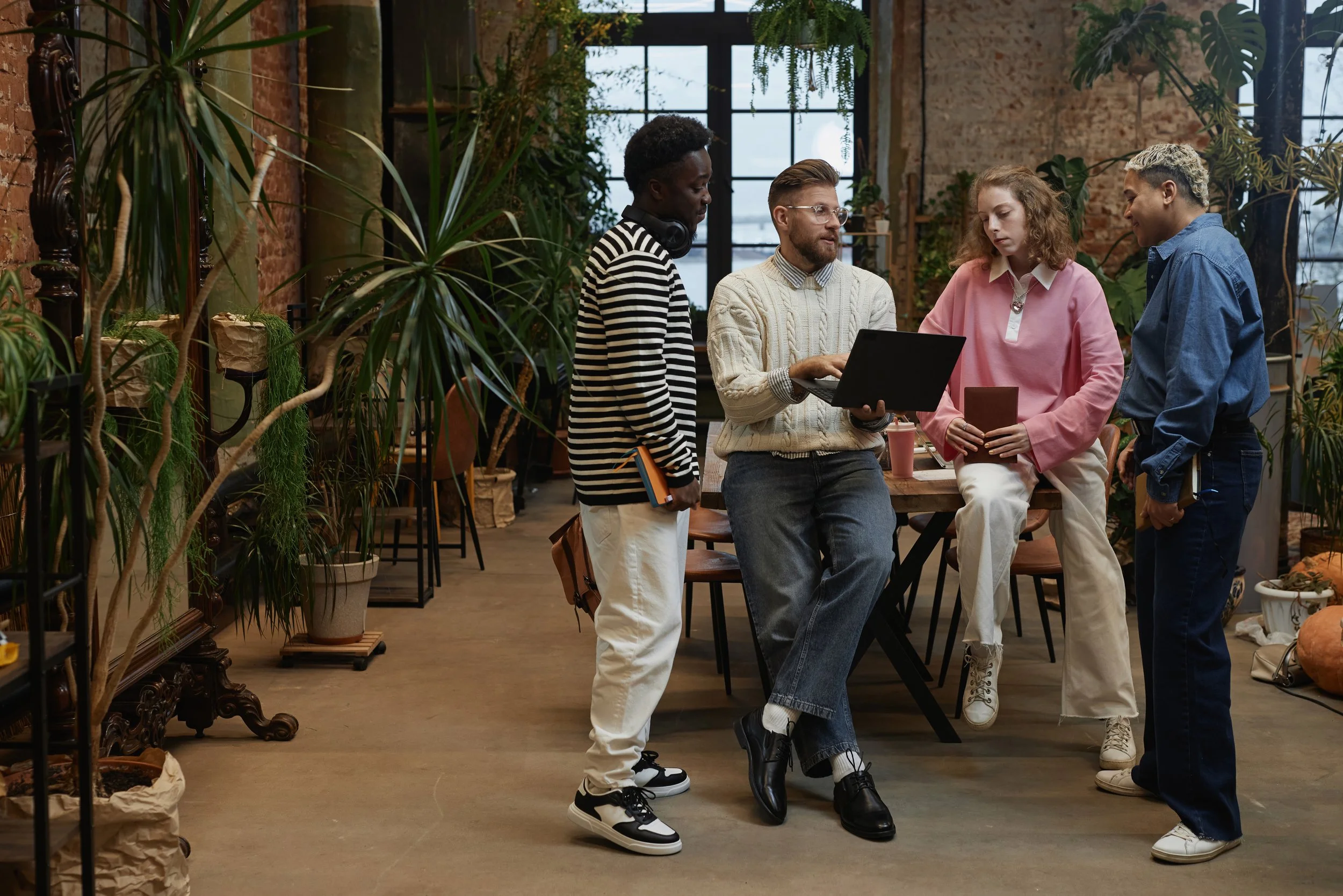 Four people stand and sit in a room with large windows and indoor plants, engaged in conversation. The room has exposed brick walls and a rustic atmosphere.