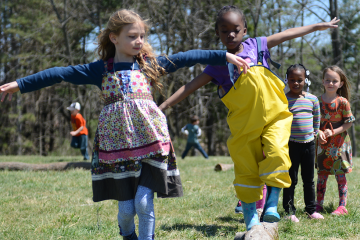 Children playing outdoors, with two girls holding hands and jumping, and other kids standing in the background on a grassy field.