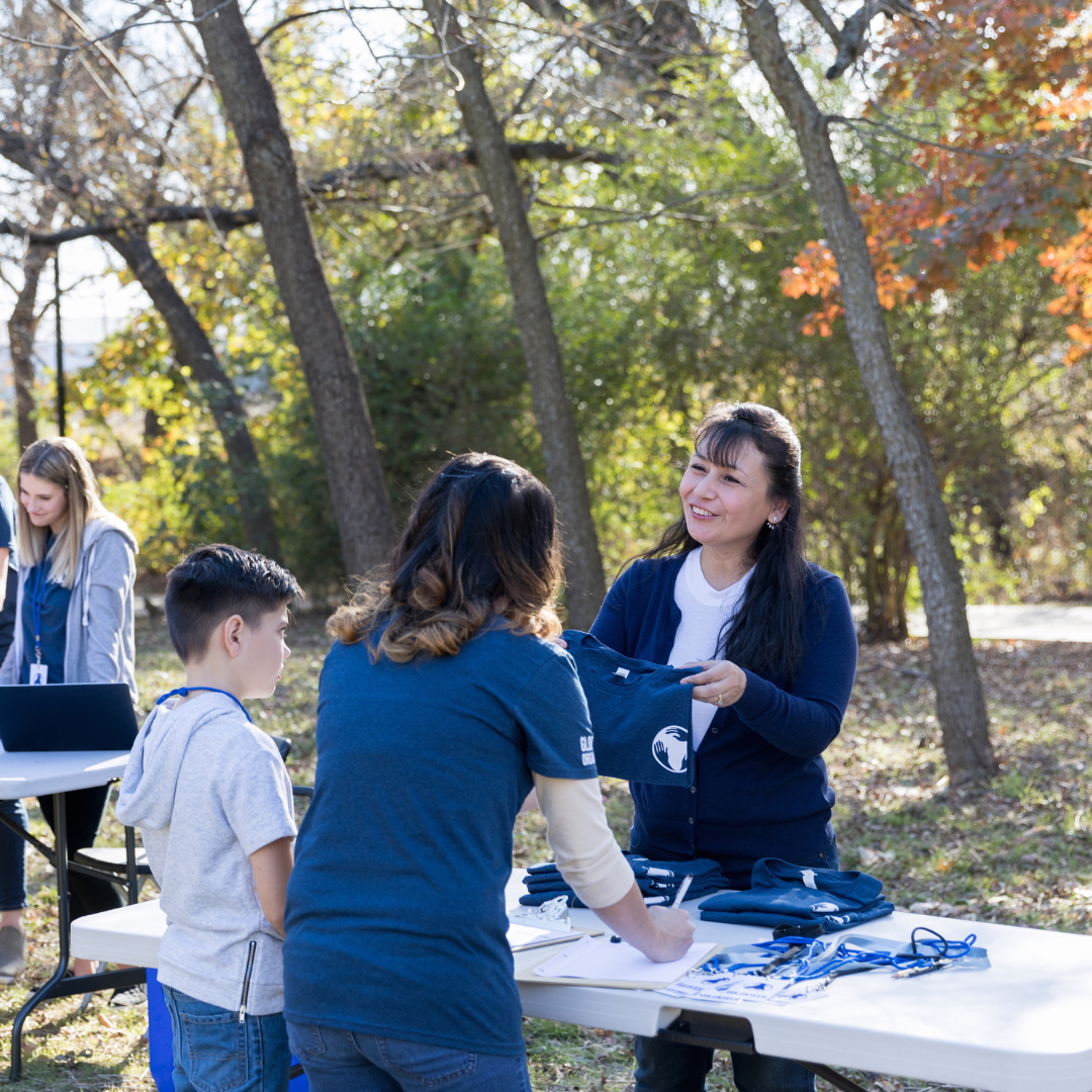 An outdoors event with children and a woman at a table surrounded by trees. The woman is handing a navy blue shirt to a girl, and there are various items on the table including lanyards and paper.