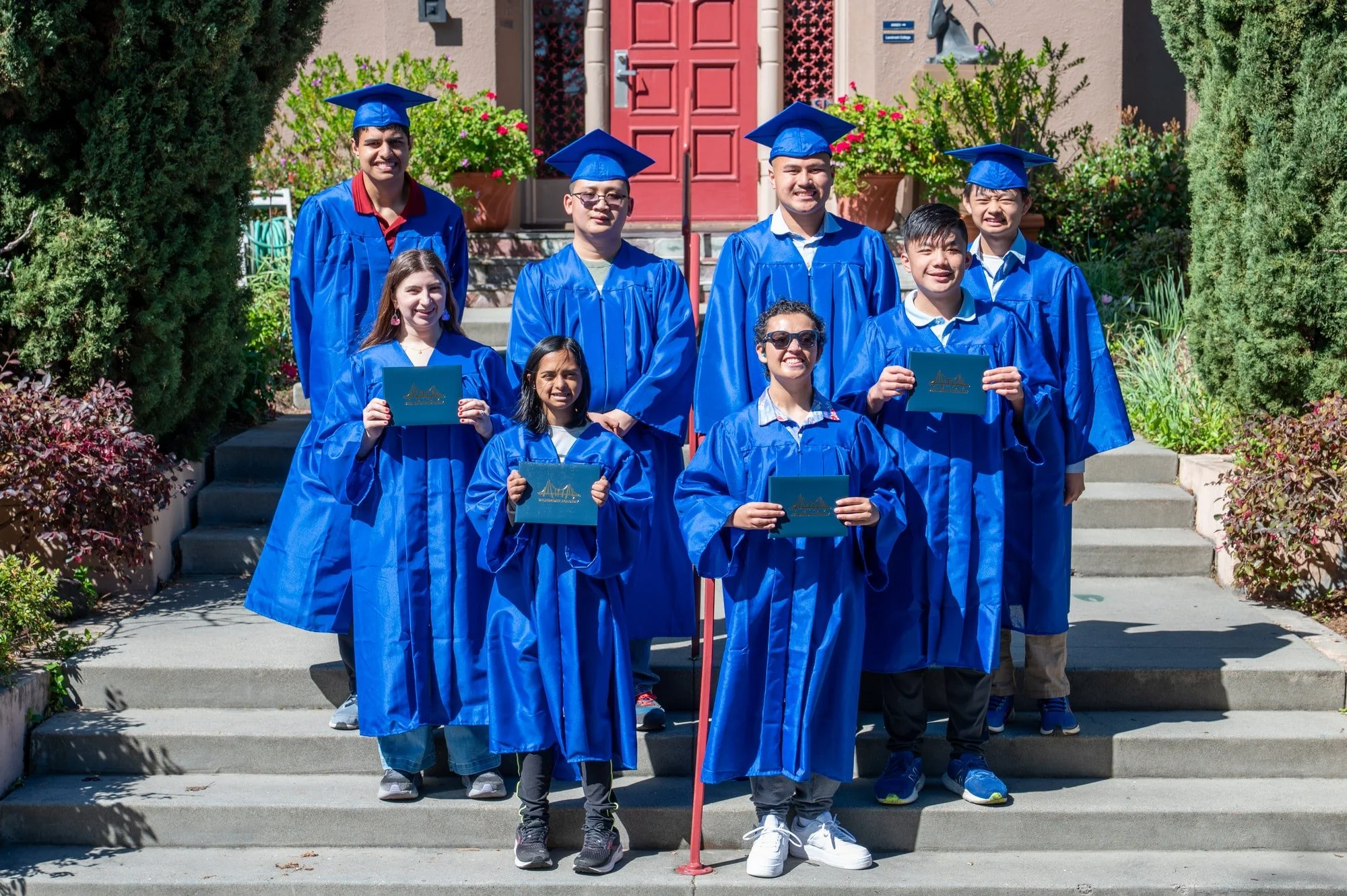 Group of eight diverse students in blue graduation gowns and caps standing on steps outside a house, holding diplomas, celebrating graduation.