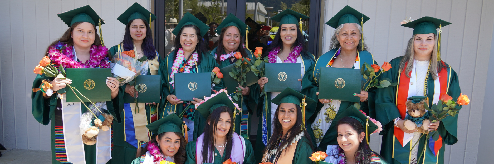 Group of women wearing green graduation caps and gowns, holding diplomas and flowers, celebrating graduation outside a building.