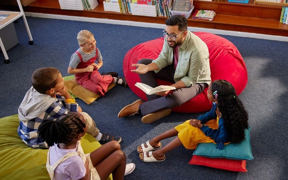 A man reading a storybook to a group of children seated on colorful cushions in a library.