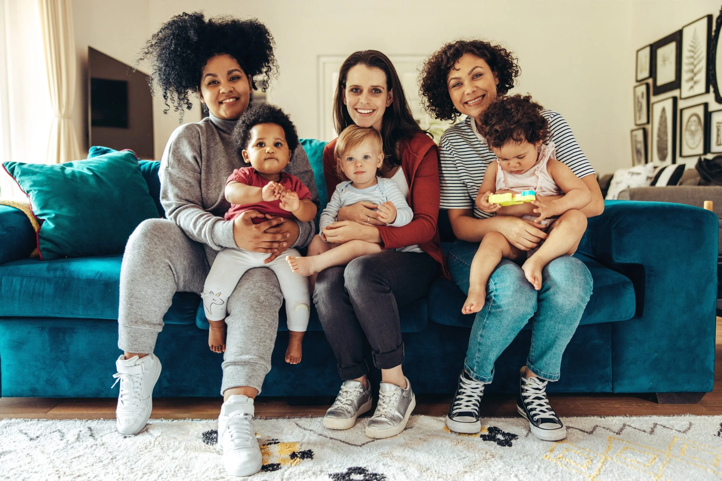 Group of six women, including three adults and three children, sitting together on a teal sofa in a living room, smiling at the camera.