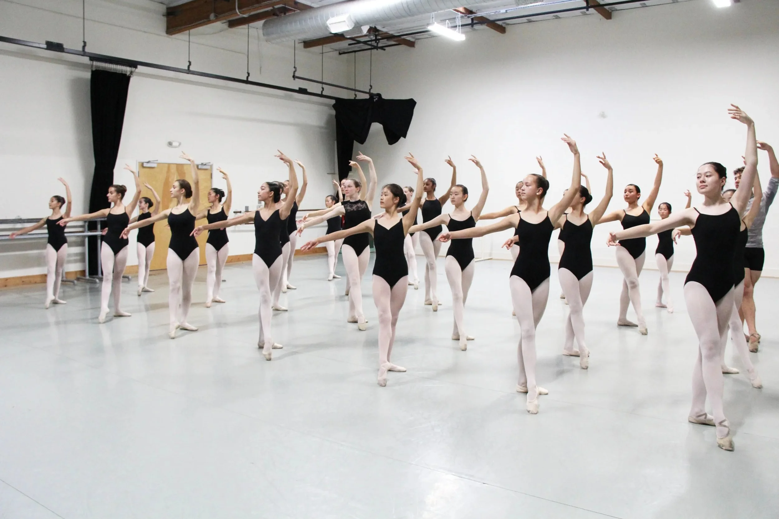 Group of ballet dancers practicing in a dance studio with white walls and wooden ceiling beams, all wearing black leotards, pink tights, and ballet slippers.