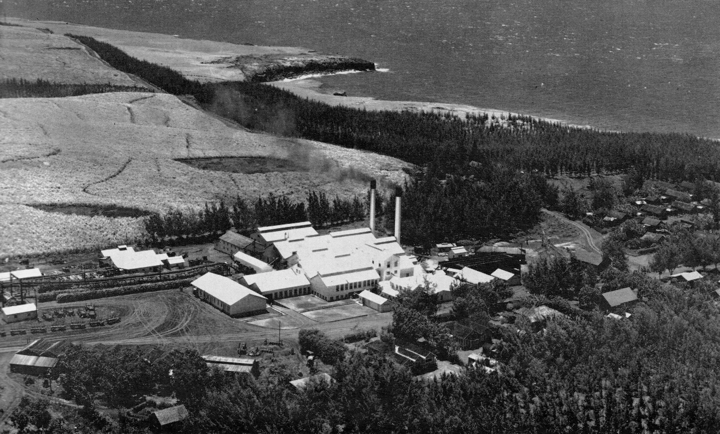 Aerial black-and-white photograph of a small town near a coastline, featuring a cluster of buildings including a factory with two tall smokestacks, houses, and fields, with trees and roads surrounding the area.