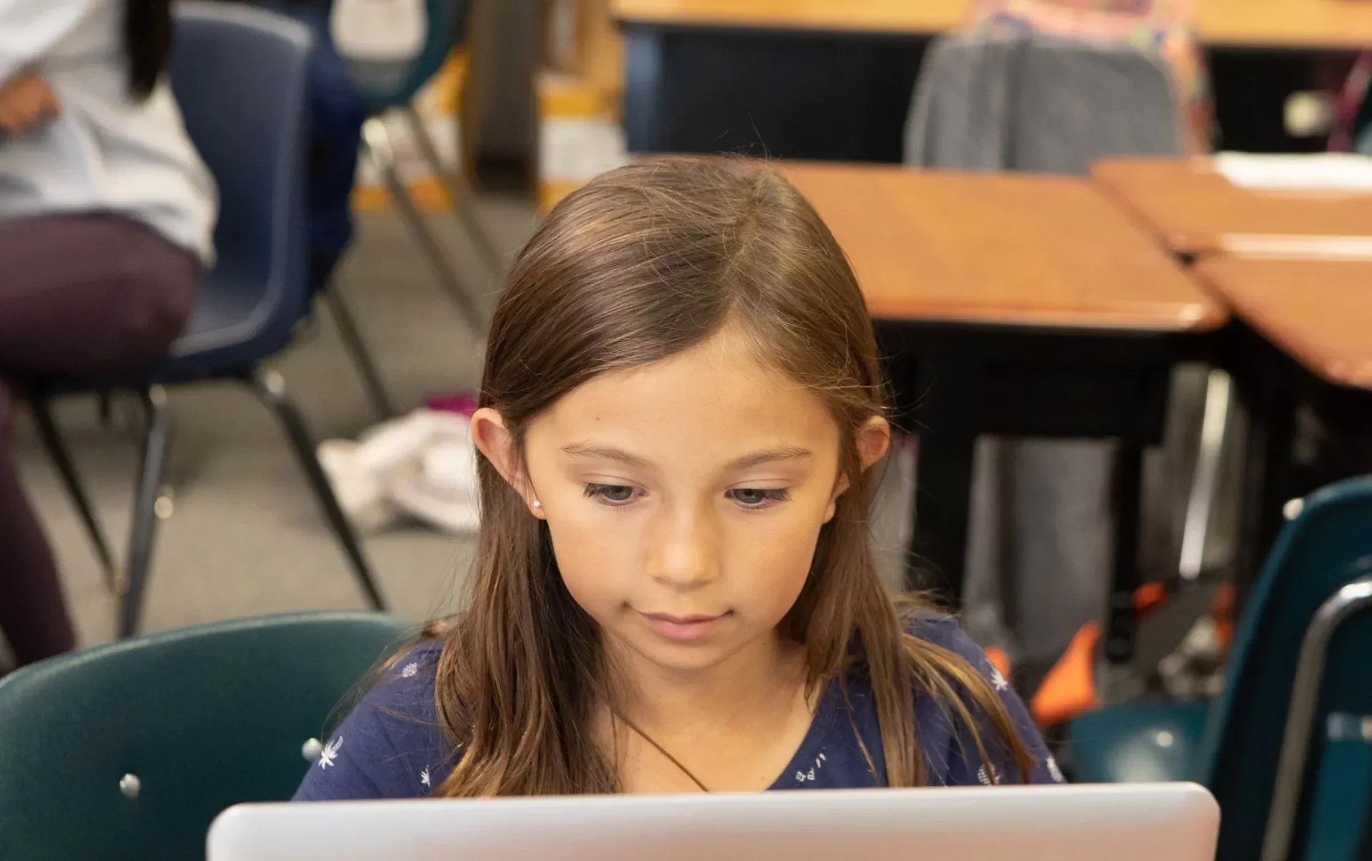 A young girl with brown hair and light skin sitting in a classroom, looking down at a tablet or device in front of her. The classroom has chairs and desks with other students in the background.