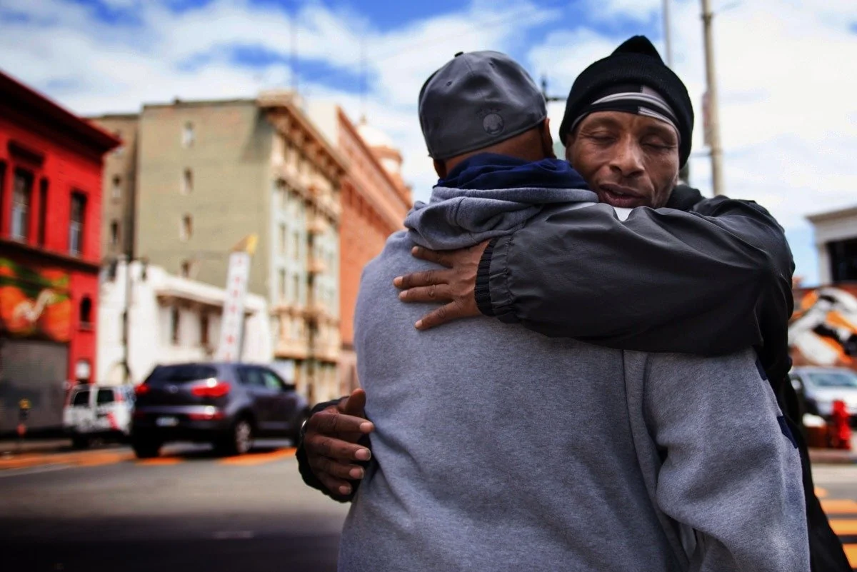 Two men embracing in a hug on a city street with colorful buildings and parked cars in the background.