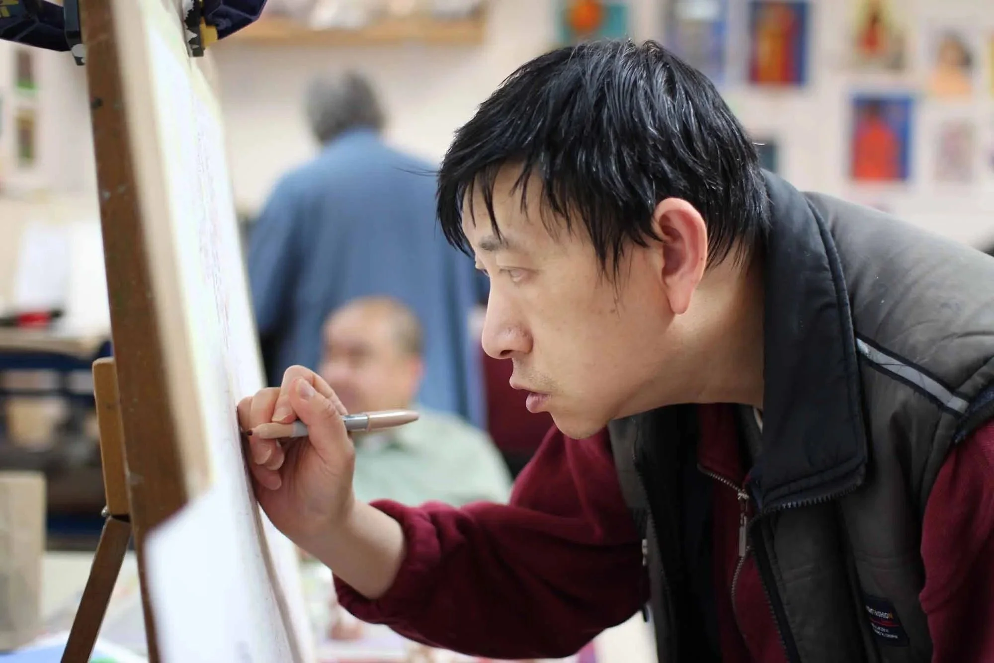 A young man with black hair writing on a whiteboard in a classroom or workshop setting, with other students in the background.