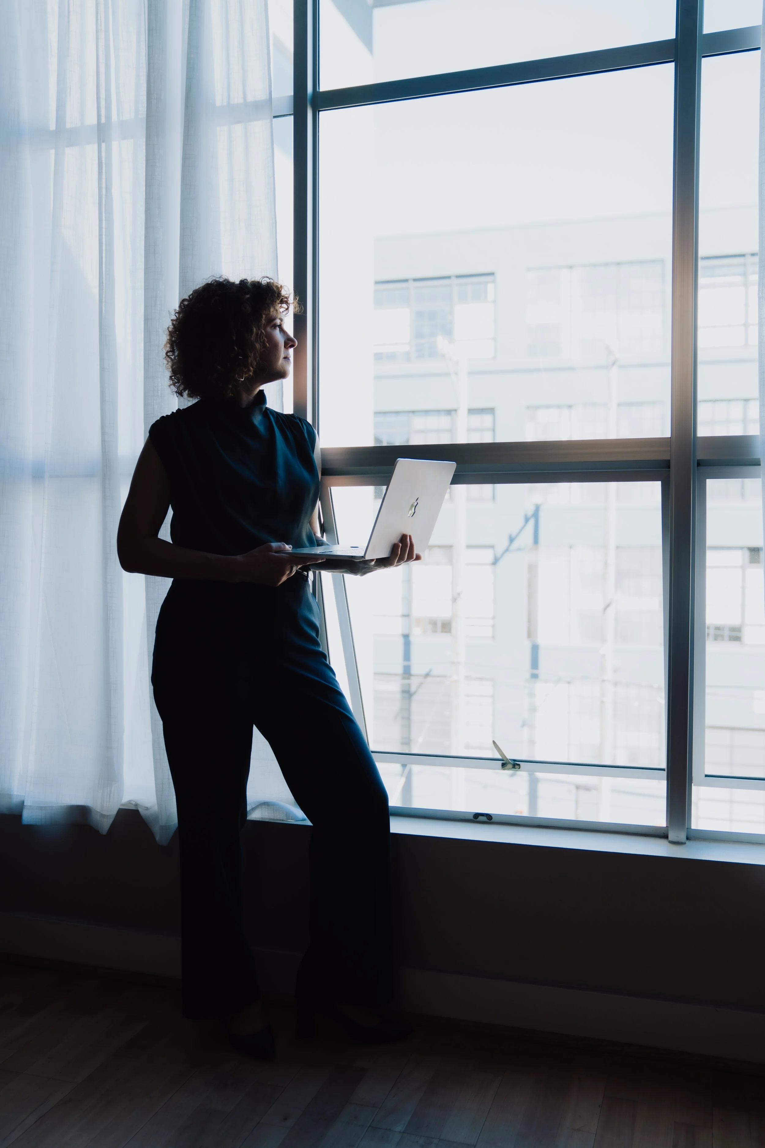 A woman stands by a large window holding a silver laptop, looking outside. The room has sheer curtains.