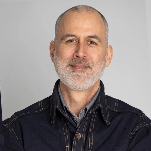 Portrait of a man with short gray hair and a beard, wearing a dark denim shirt, smiling against a plain gray background.