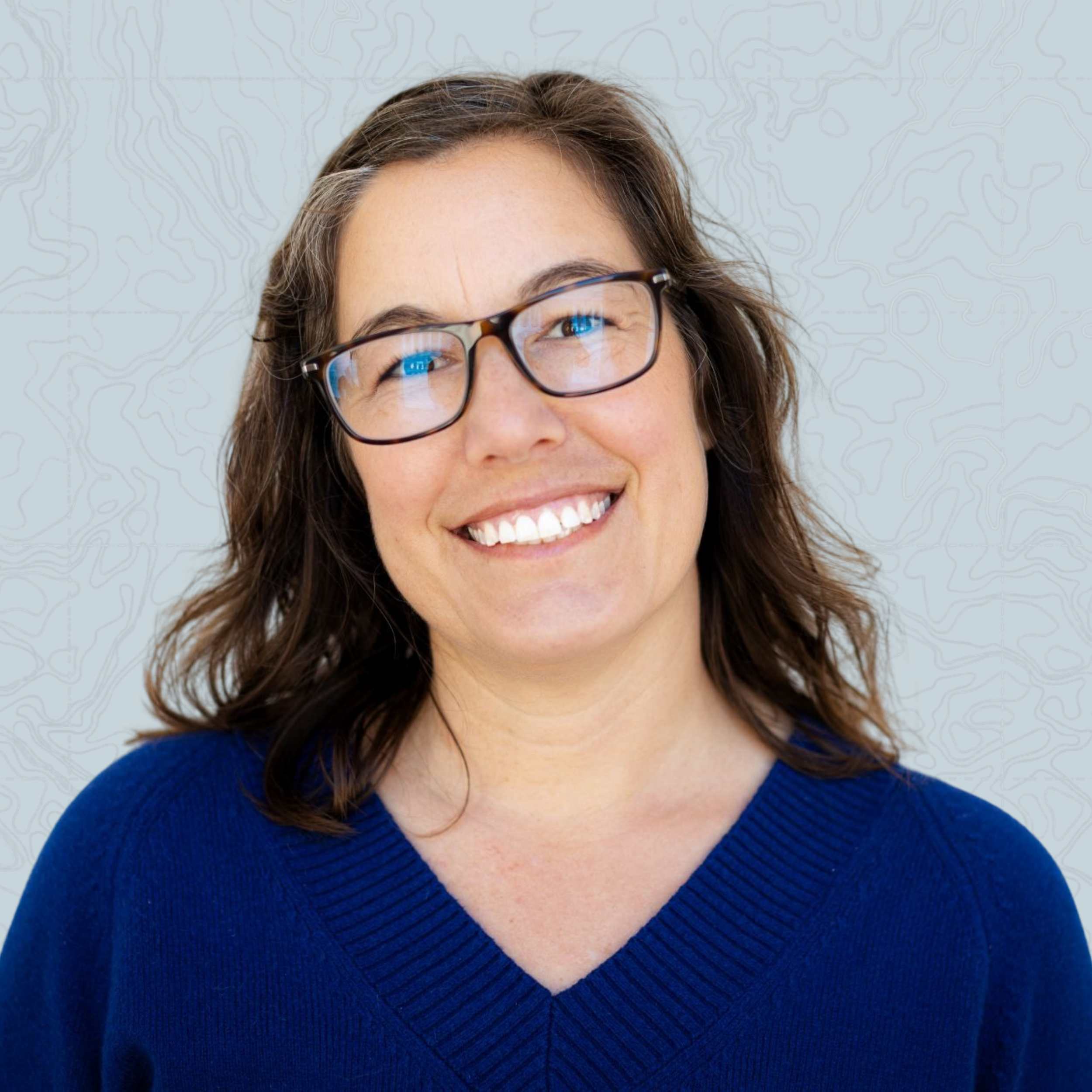 Close-up of a woman with brown hair, wearing glasses and a blue sweater, smiling.