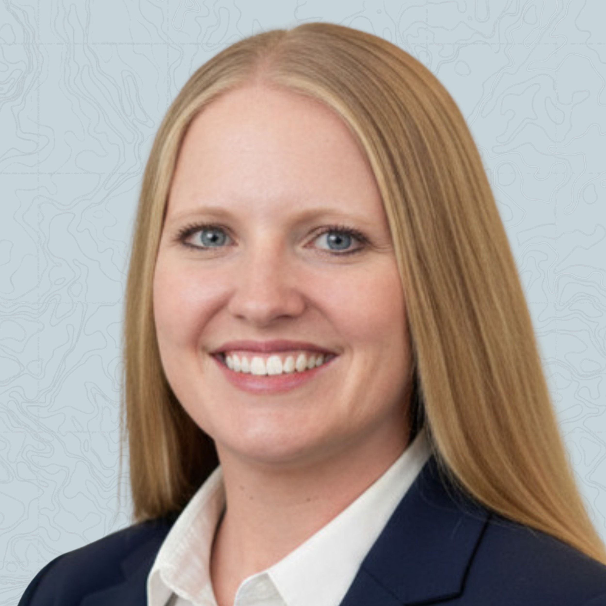 Close-up of a smiling woman with blonde hair, blue eyes, wearing a black blazer and white shirt, standing against a light, textured background.