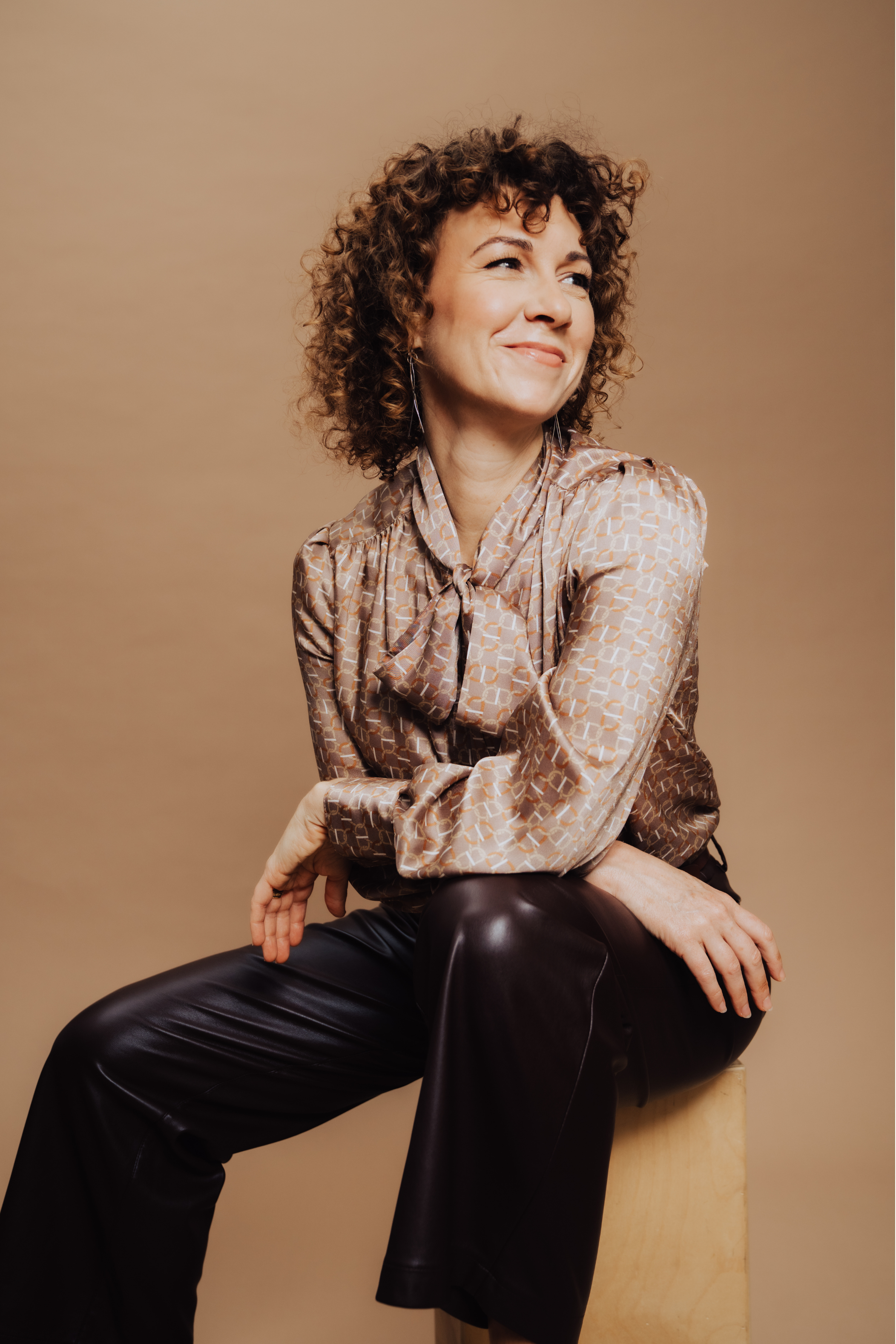 A woman with short, curly hair sitting on a wooden stool against a beige background, wearing a patterned long-sleeve blouse and black leather pants, smiling thoughtfully with her arms crossed