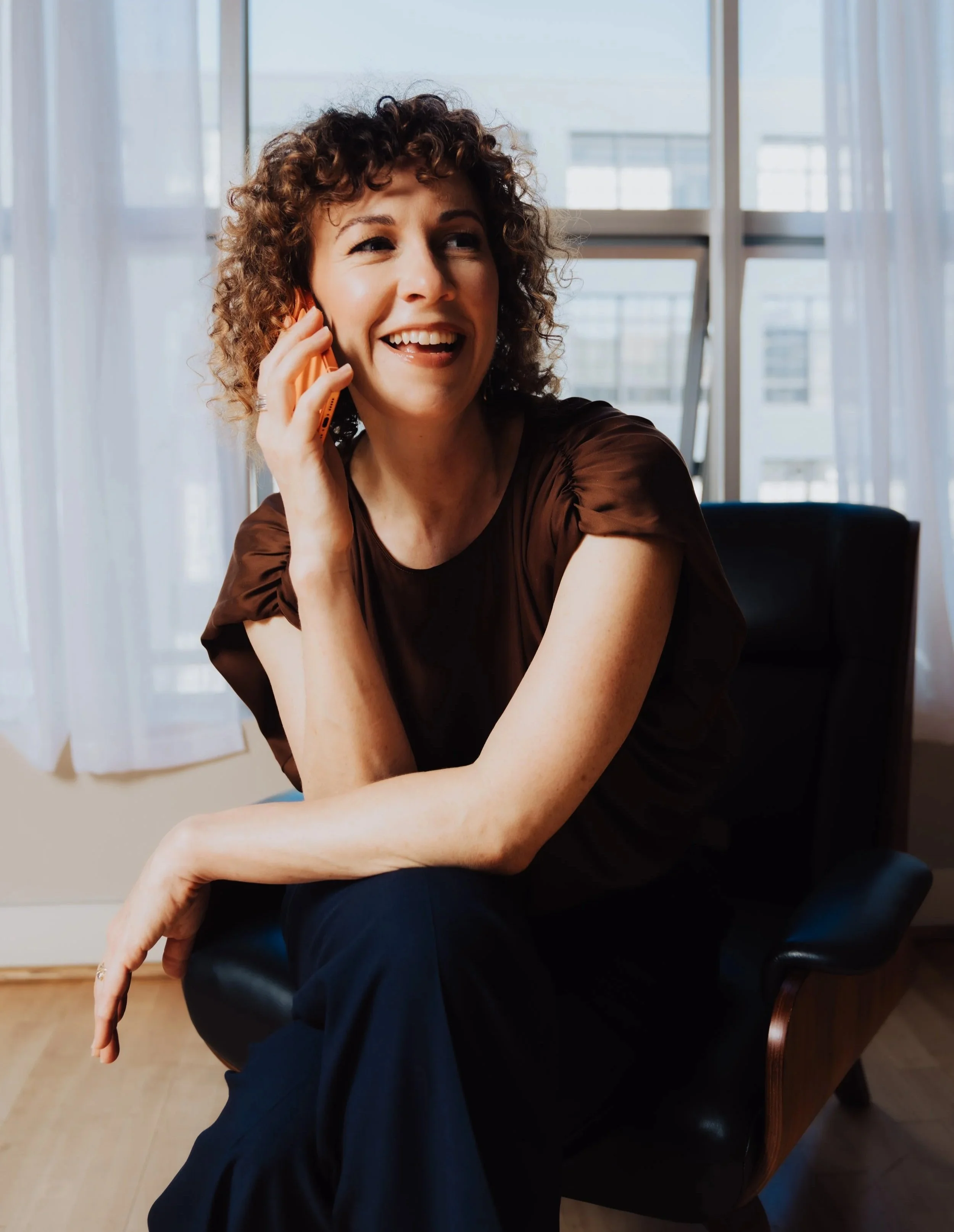 A woman with curly hair smiling while talking on a phone, sitting in a room with large windows, sheer curtains, and a wooden floor.