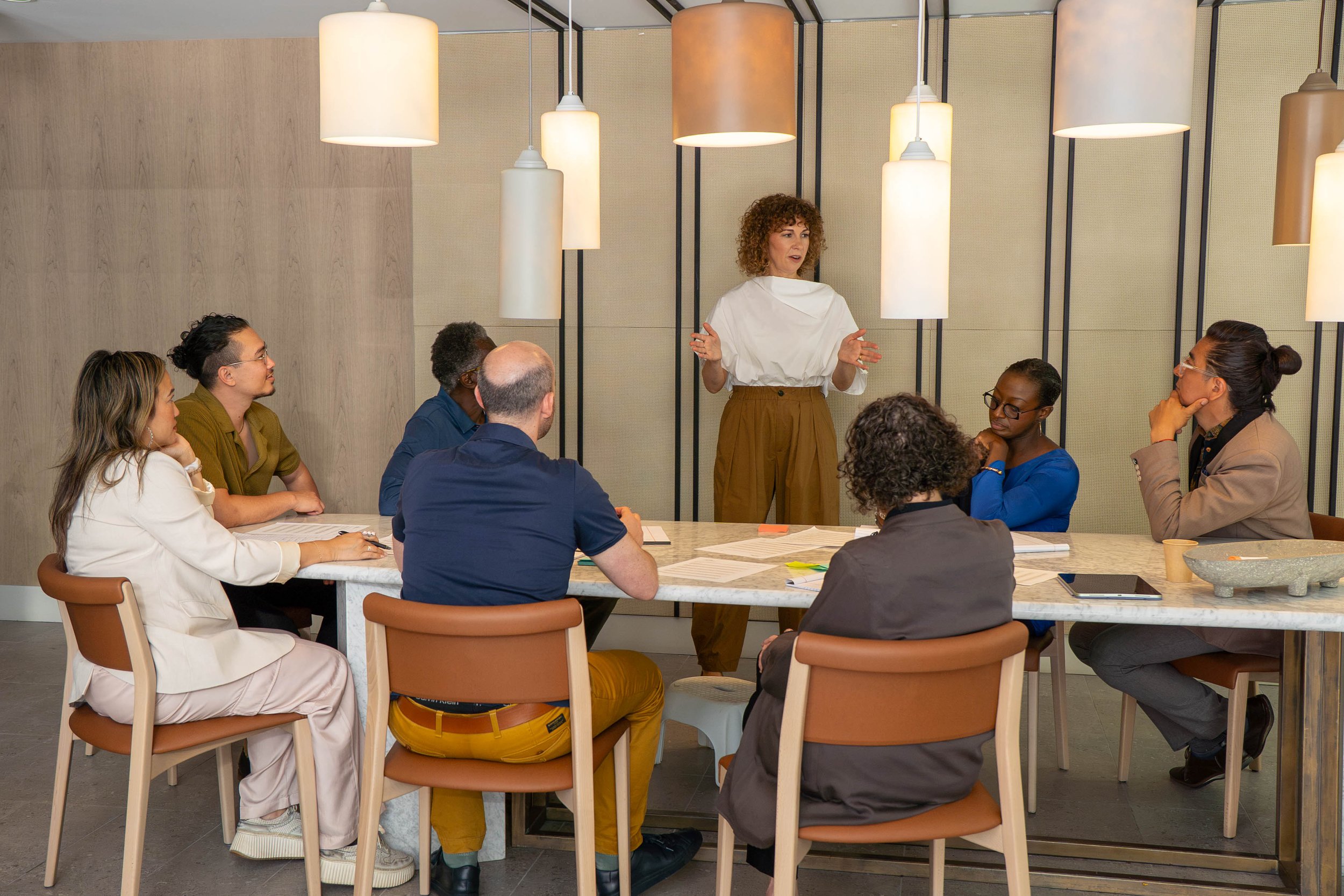 A woman standing and speaking to a group seated around a table in a modern, brightly lit room with hanging pendant lights.
