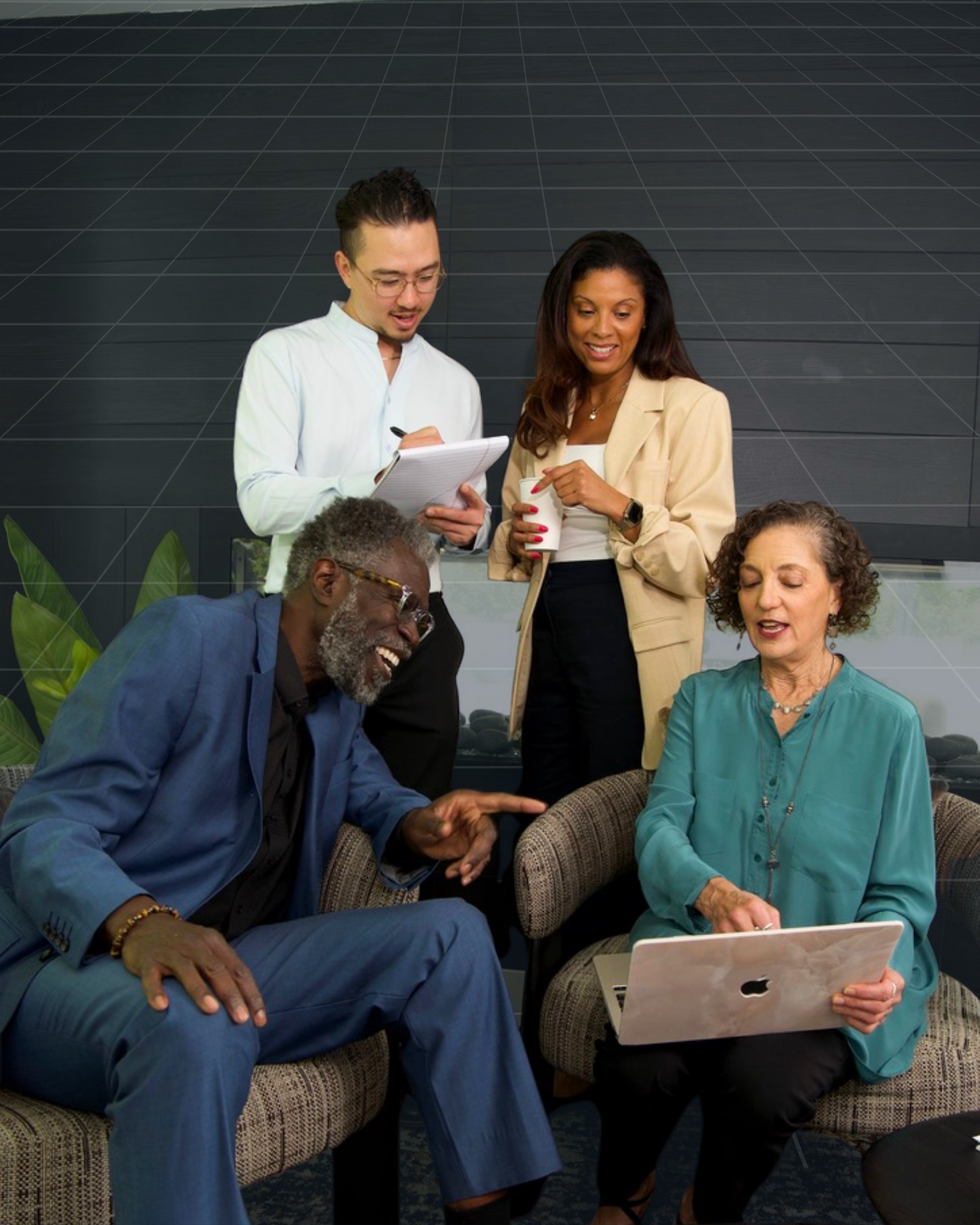 Group of five diverse adults engaging in a conversation in a modern office setting. Two people are sitting on a sofa, one man is smiling and gesturing, while a woman is using a laptop. Two others are standing nearby, one woman holding a coffee cup and looking at the laptop.