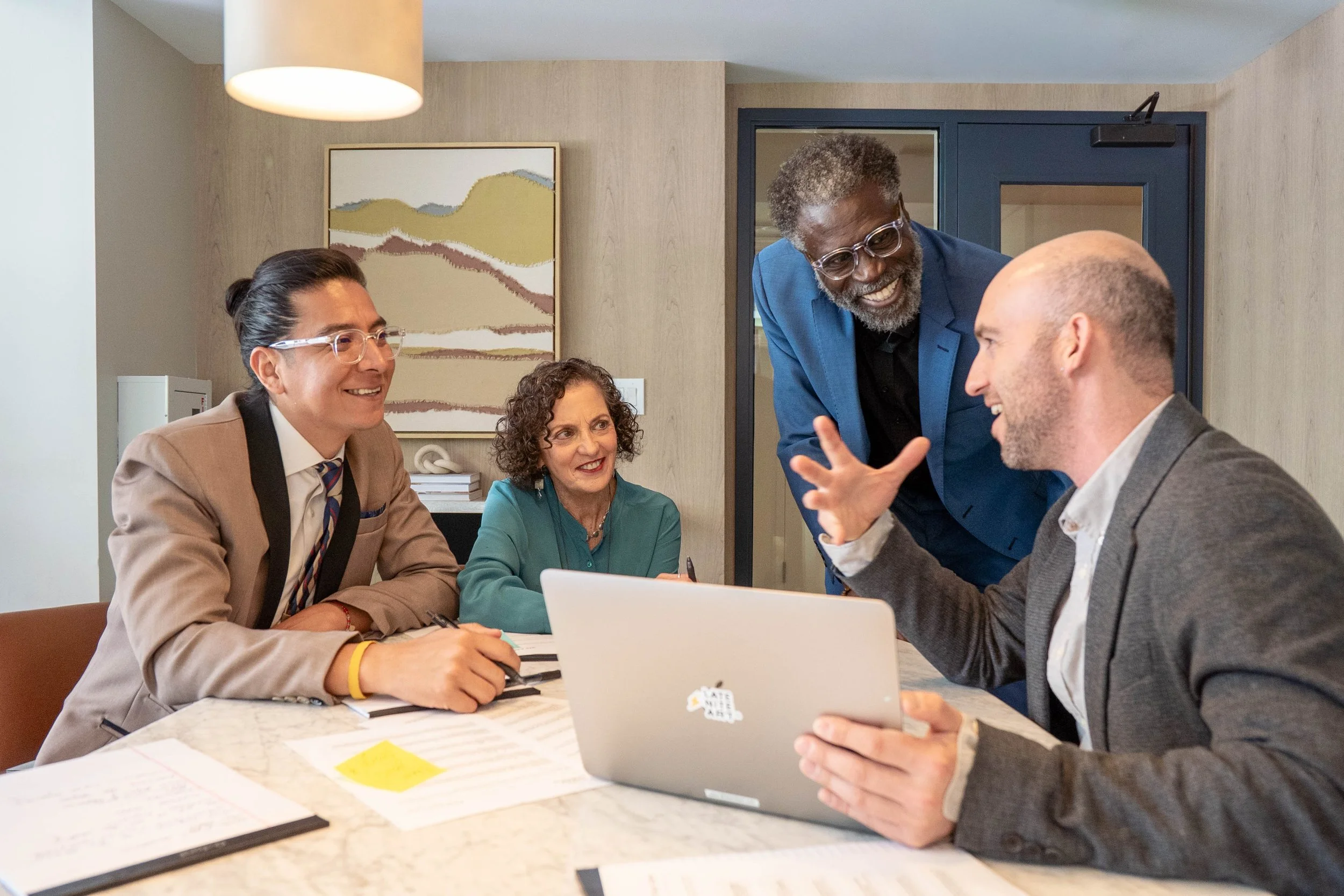 A diverse group of four professionals engaged in a lively discussion at a meeting, with one person using a laptop.