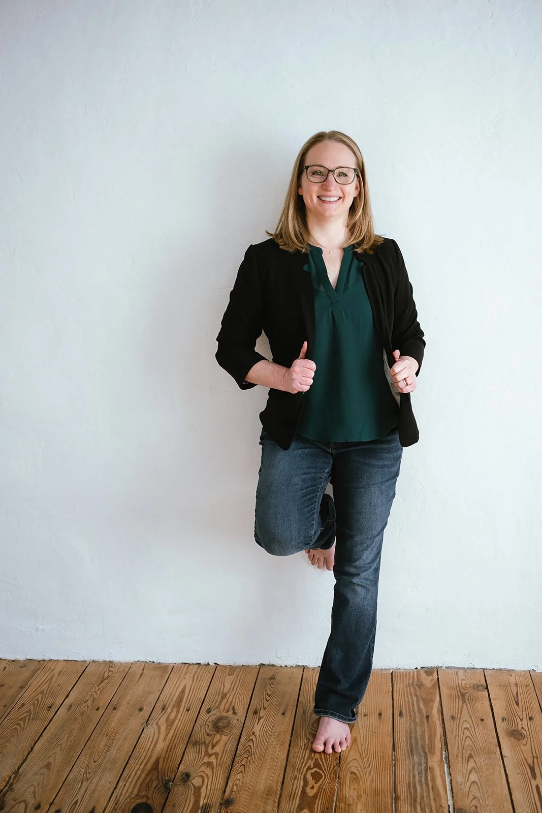 Dr Amanda Townsend wearing a black blazer over a dark green blouse and jeans, standing barefoot on a wooden floor against a plain white wall.