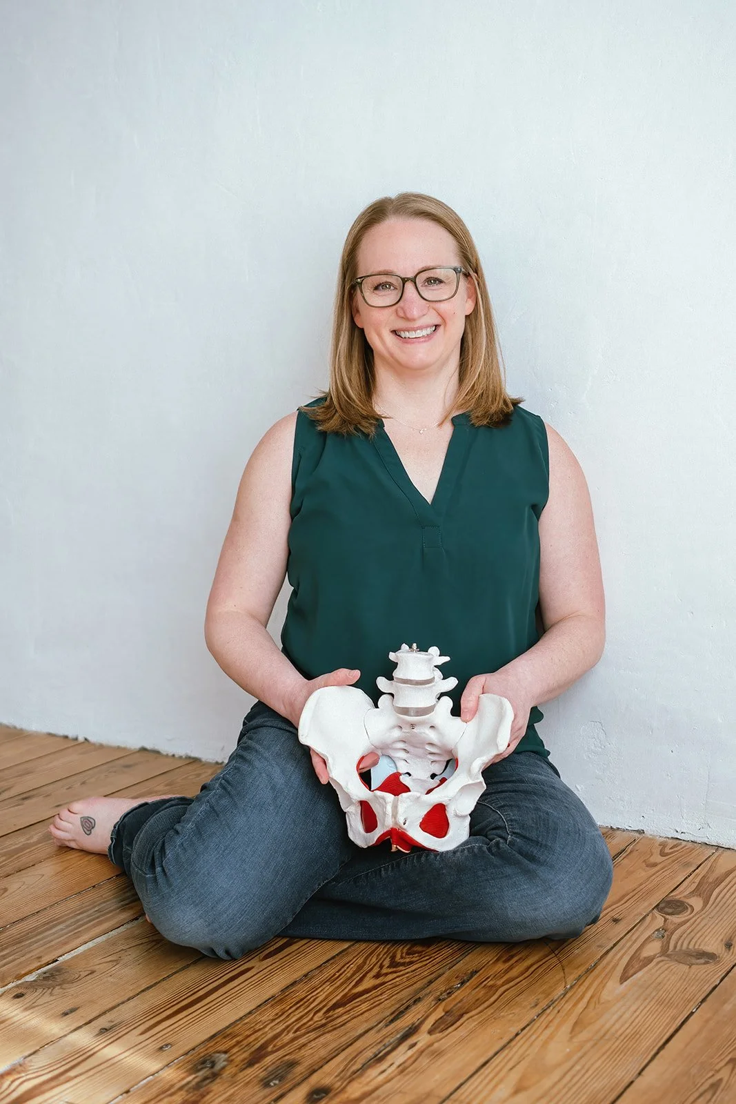 Woman with glasses sitting on wooden floor holding a pelvis and lower spine model, smiling, wearing a sleeveless dark green top.