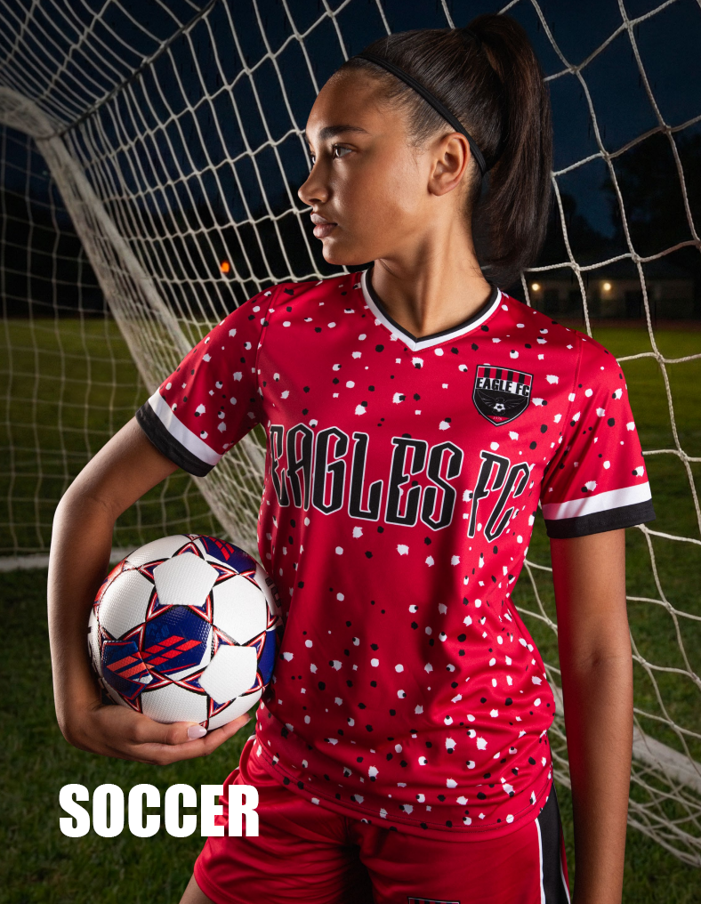 A young female soccer player standing by a goal net on a soccer field during evening, holding a soccer ball, wearing a red jersey and a logo, and red shorts.