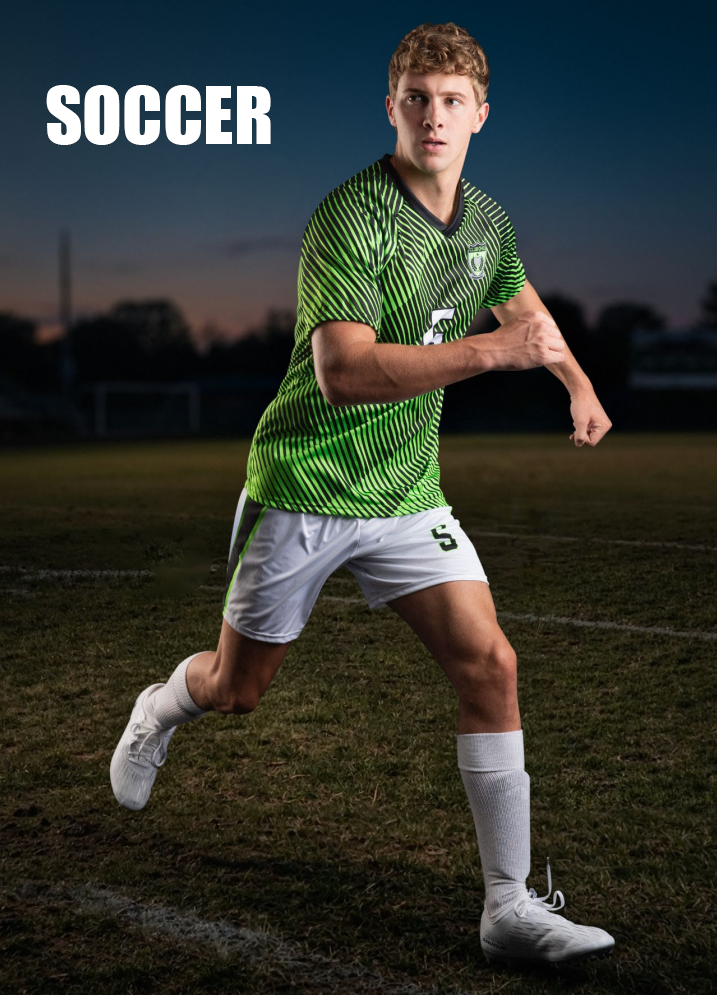 A young male soccer player in a green and black striped jersey and white shorts runs on a soccer field during dusk.