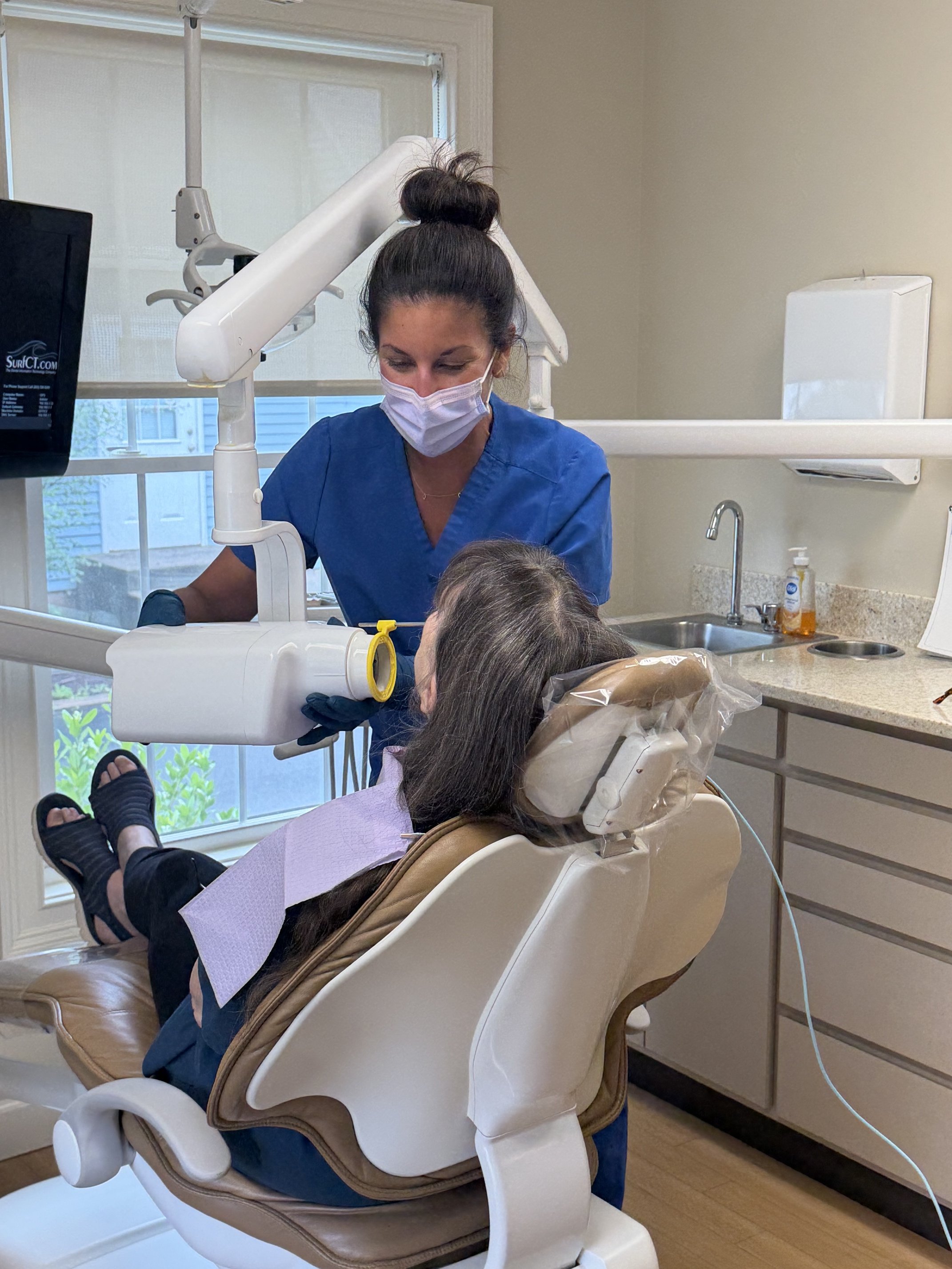 A dental professional wearing a face mask and blue scrubs working on a patient in a dental clinic. The patient is seated in a dental chair with a dental light overhead and a window in the background.