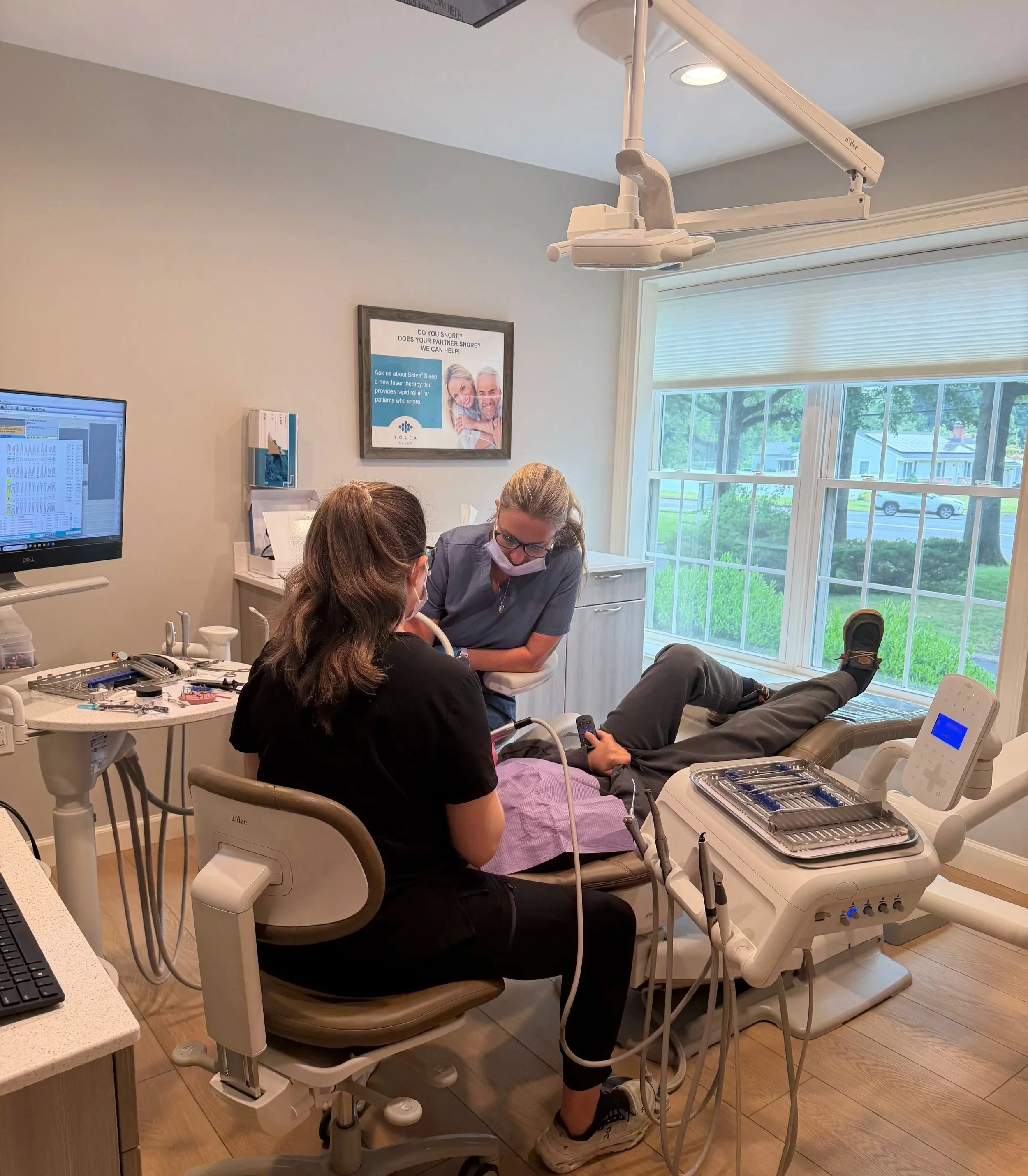 A dental clinic scene with a patient lying back in a dental chair, which is being attended to by a female dentist wearing scrubs, gloves, and a mask. There are dental tools and equipment on nearby trays, and a computer monitor is visible on the left side. The room has a large window with blinds, allowing natural light, and a framed poster on the wall.