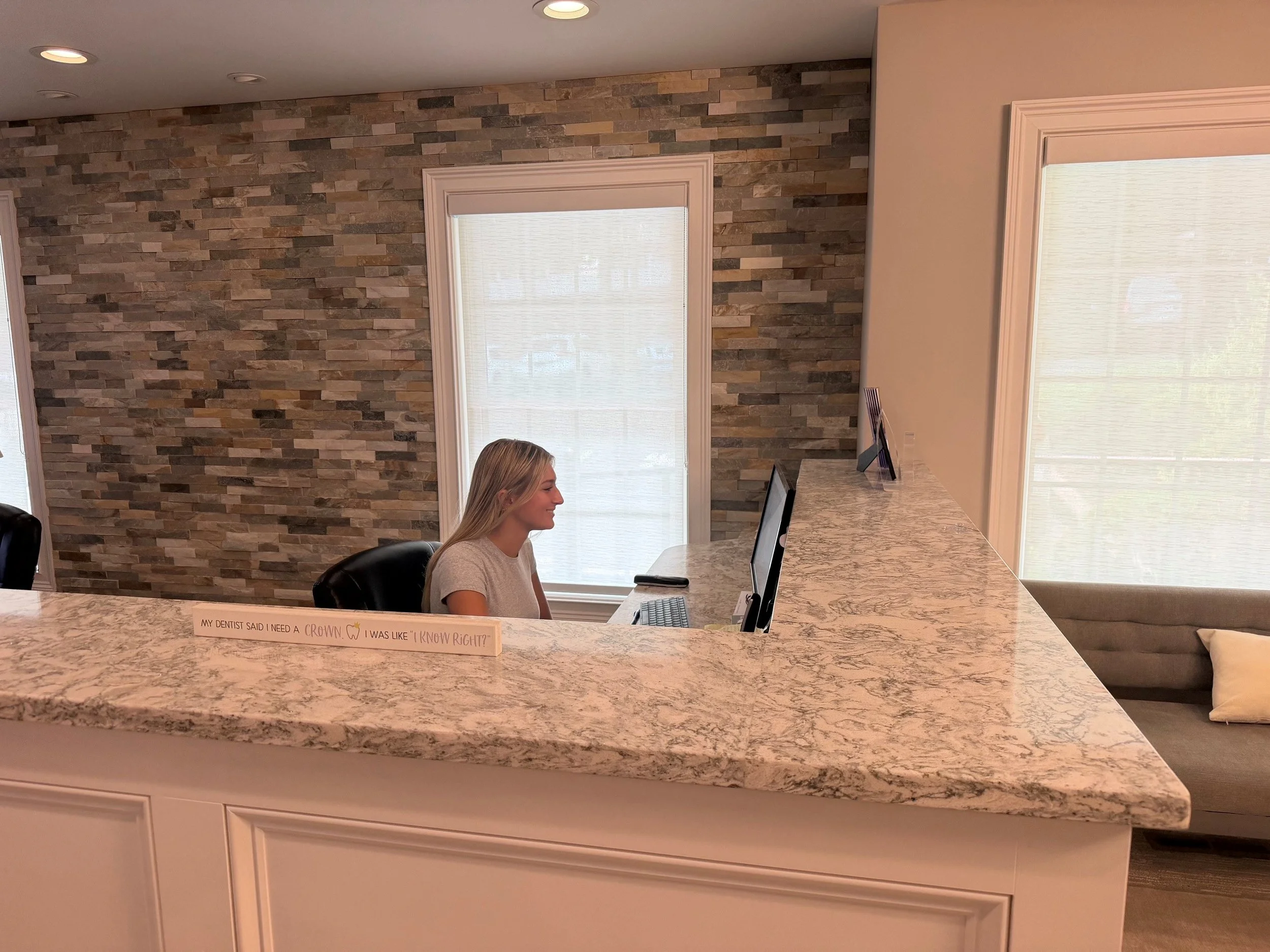 A woman sitting at a reception desk with a Neal's Yard Remedies sign in front of her. Behind her is a stone accent wall with two windows and blinds. The desk has a marble countertop and a computer monitor.