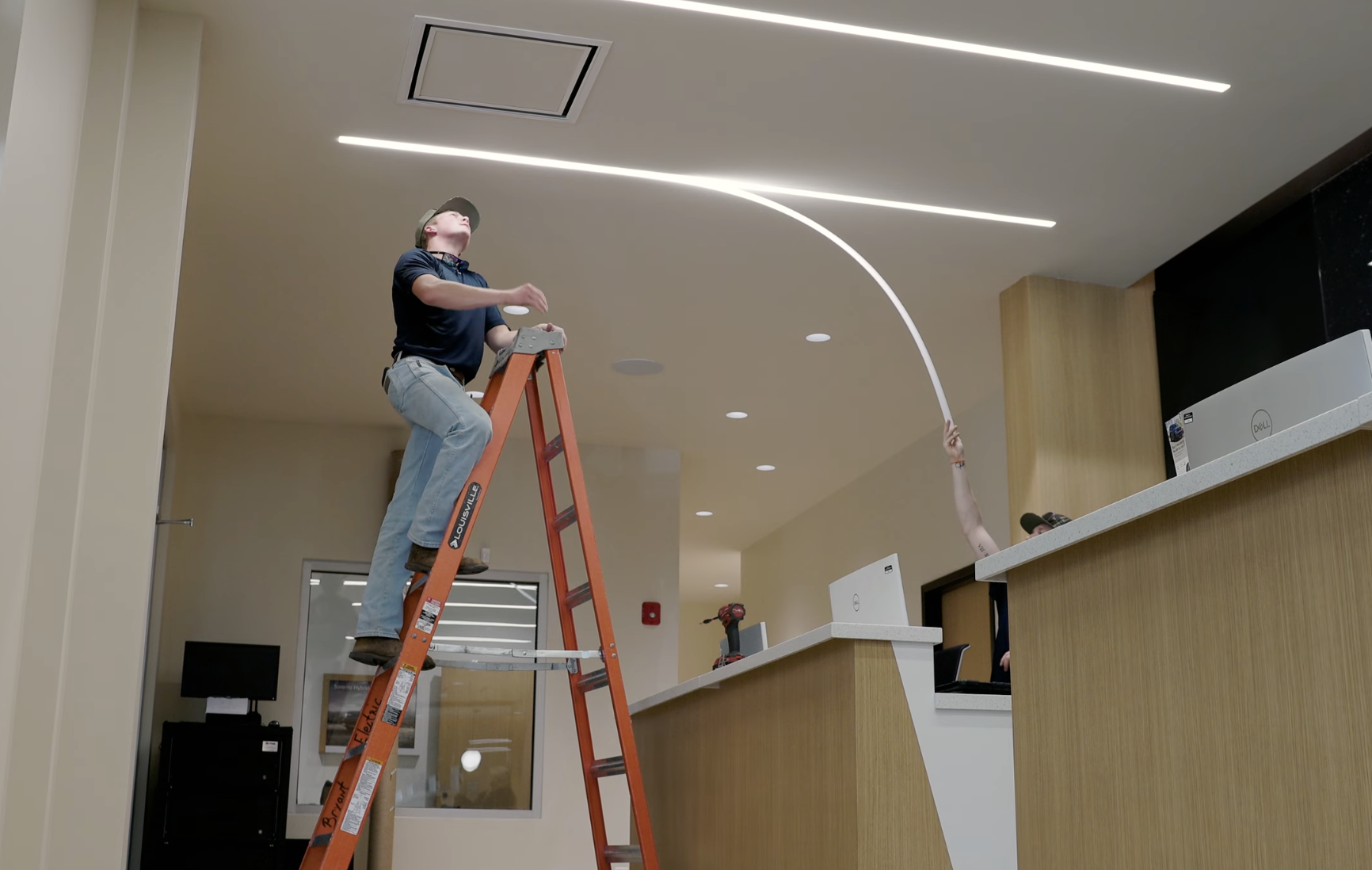 Two people working on installing or repairing LED light strips on a ceiling. One person is elevated on an orange ladder, and another person's arm is visible reaching above a reception counter.
