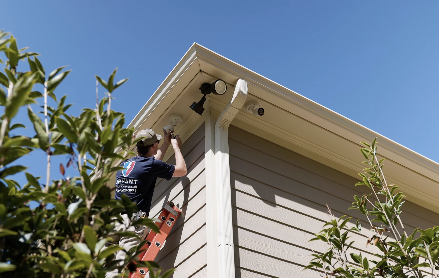 A worker on an orange ladder installing or maintaining security cameras on the corner of a house's roof. The house has beige siding, a white gutter, and a security light. The sky is blue, and there are green bushes in the foreground.