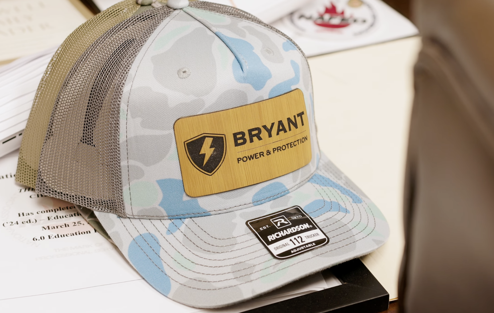 A baseball cap with a camouflage pattern and mesh back, featuring a yellow patch that says "BRYANT POWER & PROTECTION" with a black shield and lightning bolt logo. The cap is placed on a desk with papers and appears to be in an indoor setting.