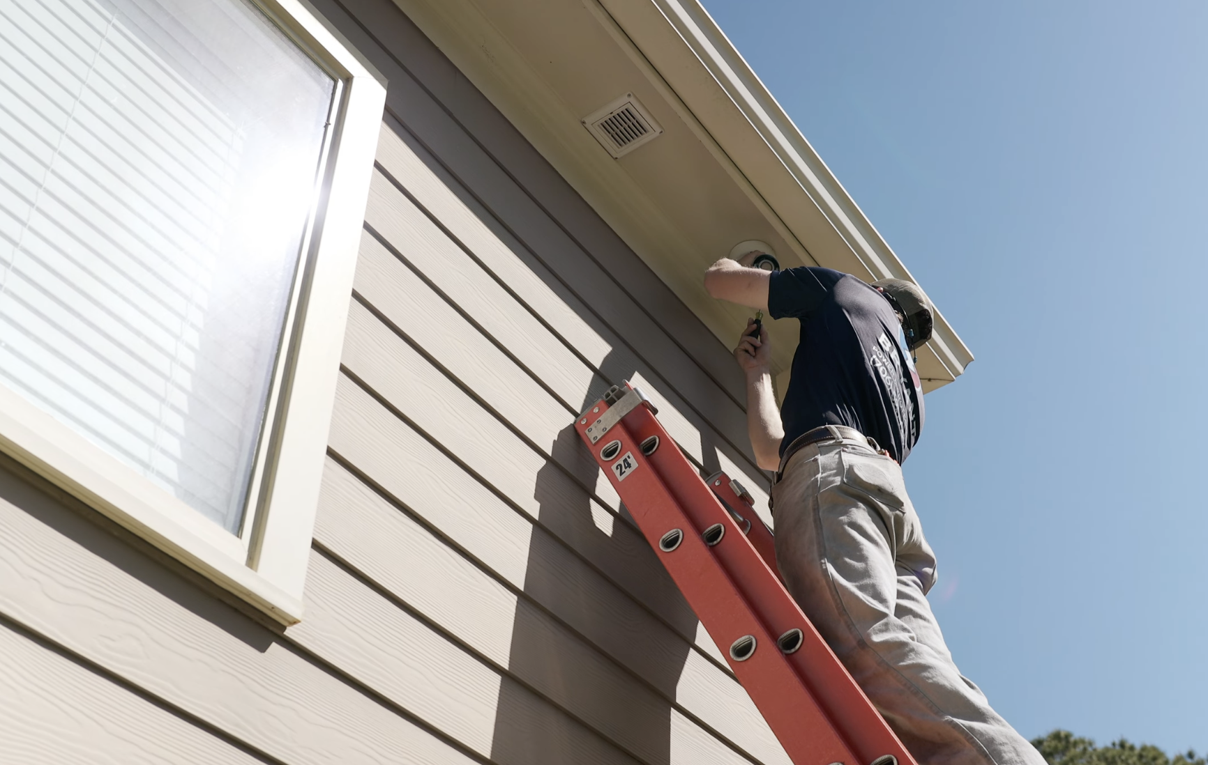 A person standing on a red ladder cleaning or inspecting the exterior siding of a house near a window under a clear blue sky.