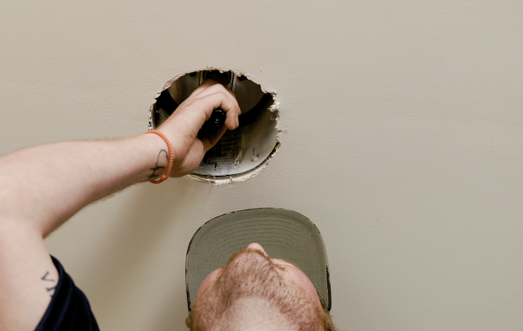 Person installing or repairing a ceiling light fixture, holding a clamp or tool with a hole in the ceiling, wearing a cap and a black shirt, with tattoos on the arm.