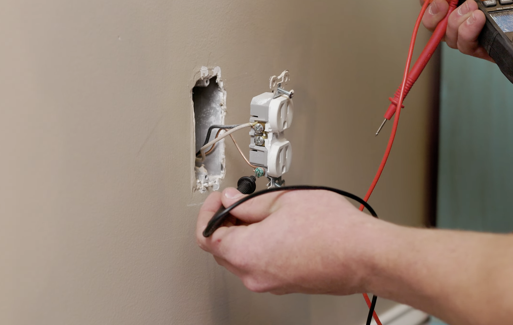 Person using a multimeter to test electrical wiring at wall outlet.
