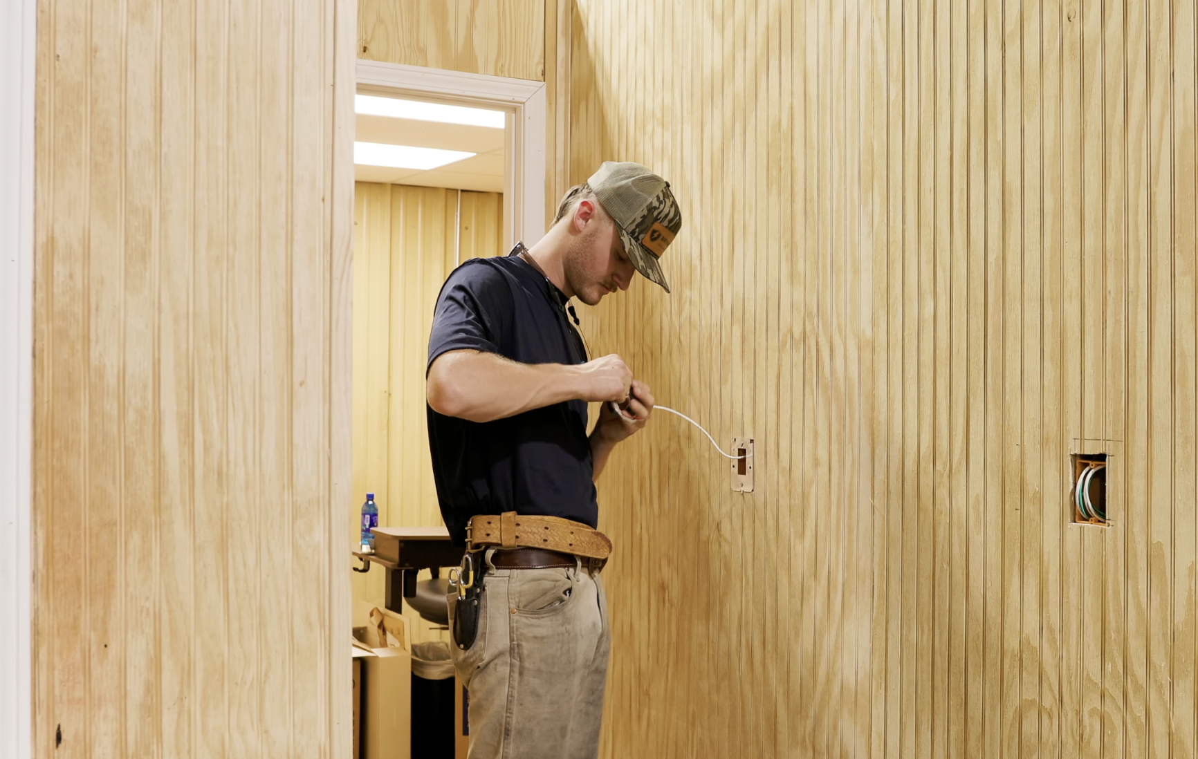 A man in work clothes, wearing a tool belt, installing or repairing electrical wiring in a room with wood-paneled walls.