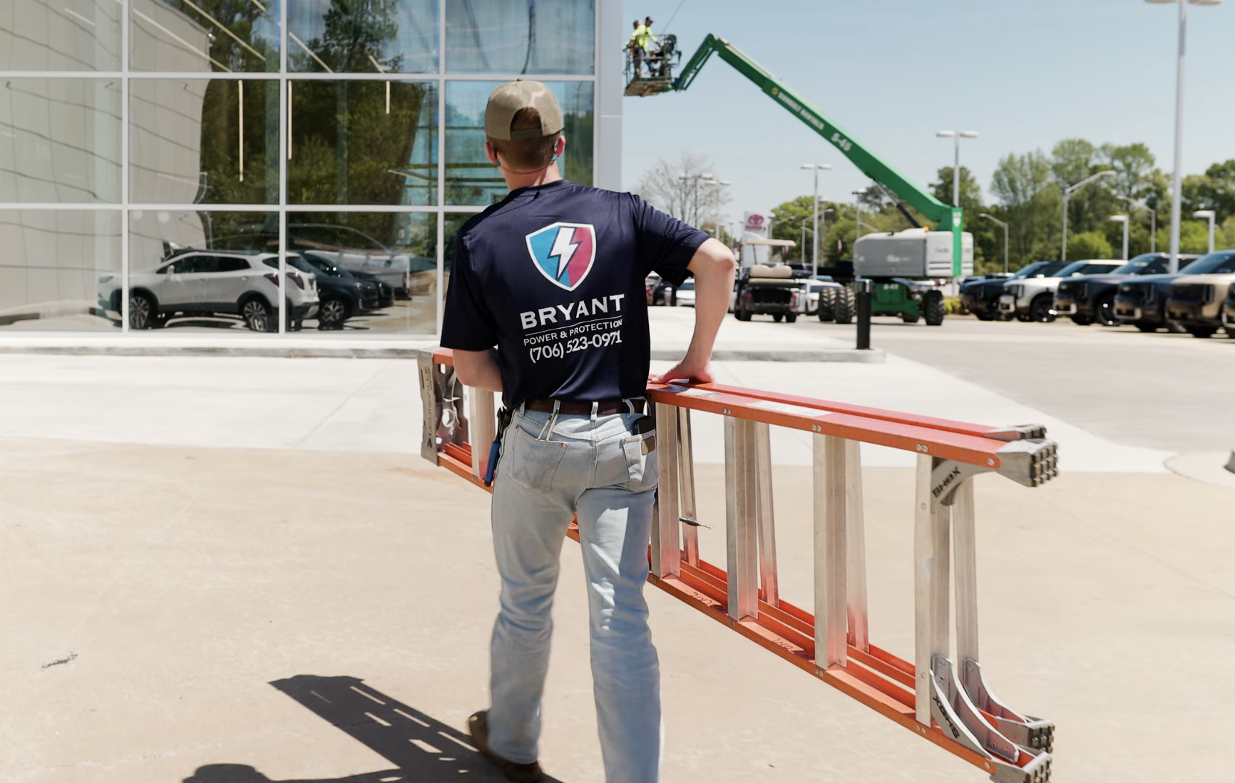 A worker wearing a navy blue shirt with the logo 'Bryant Power & Protection' is carrying an extension ladder outdoors near a commercial building with large glass windows. There are cars parked in the background, and a green lift with two workers is elevated near the building.