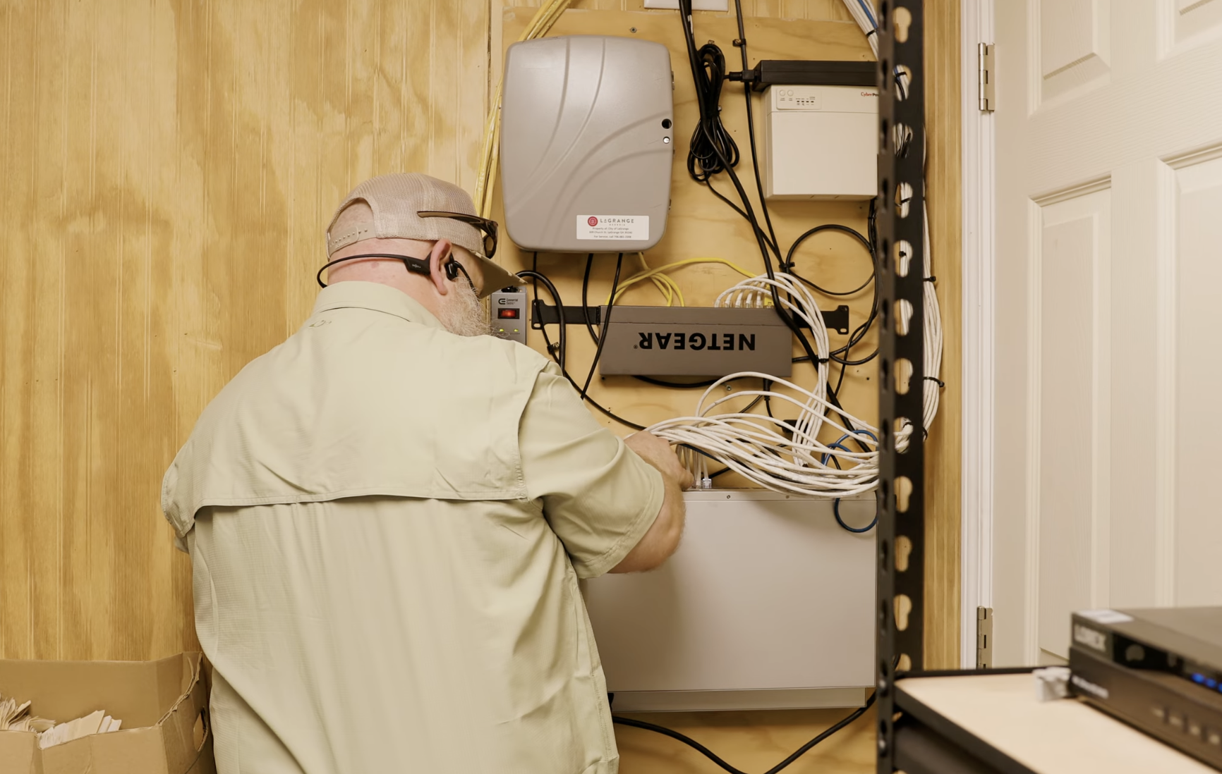 A technician wearing a beige shirt, glasses, and a tan cap working on networking equipment inside a wooden compartment, with various cables and electronic devices mounted on the wall.