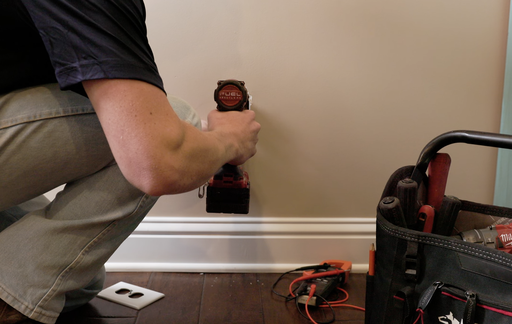 Person kneeling on the floor using a red cordless drill against a beige wall, with tools and a tool bag nearby.