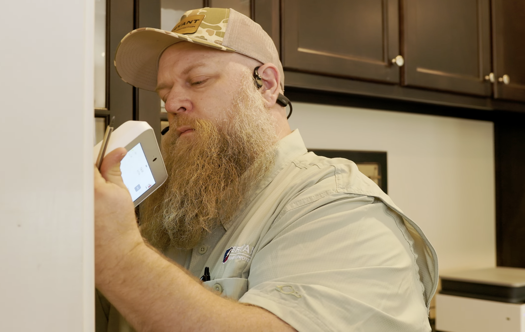 A man with a long beard, wearing a beige baseball cap and a beige shirt, is holding a white device close to his ear in a kitchen setting.