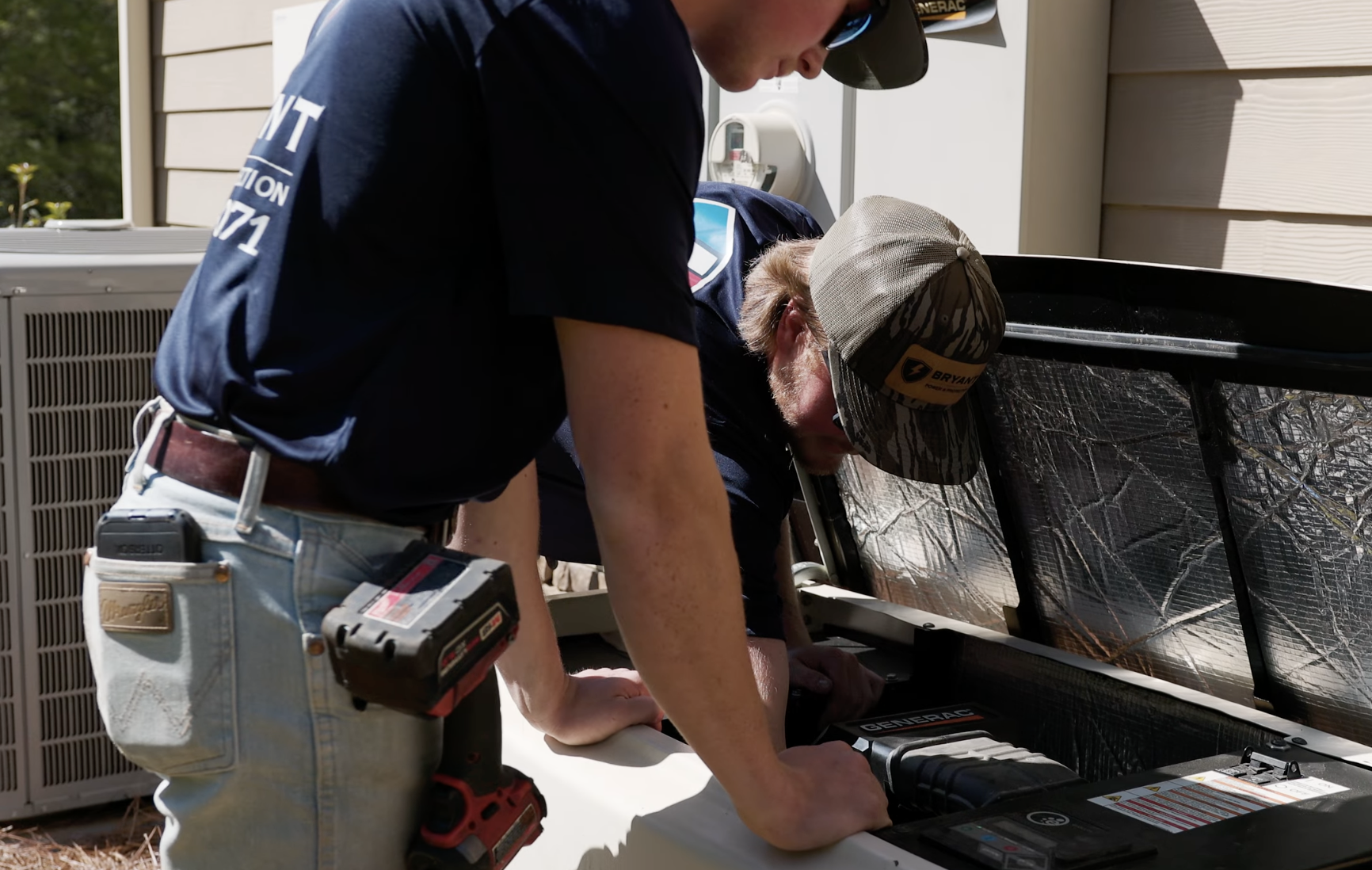 Two men are working on a generator outside of a house, with one using a power drill and the other fixing or inspecting the equipment.