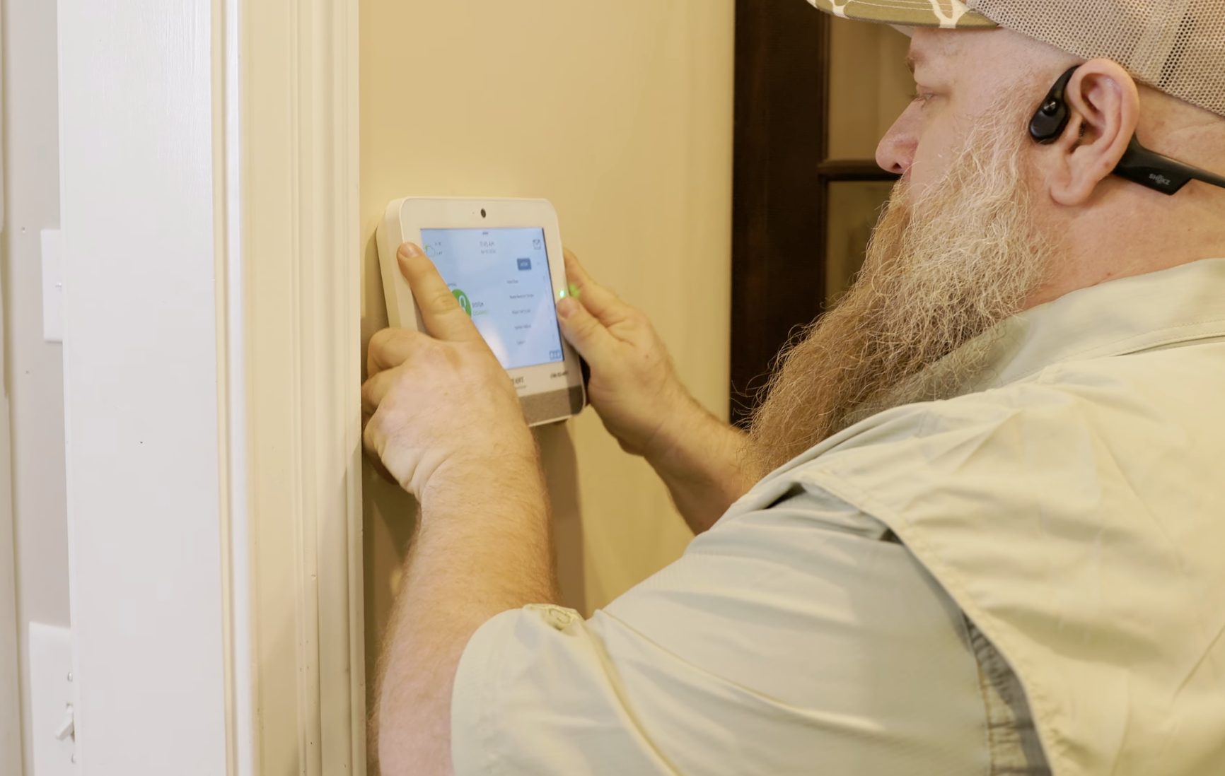 A man with a long beard wearing a beige cap and glasses is adjusting a thermostat on a wall.